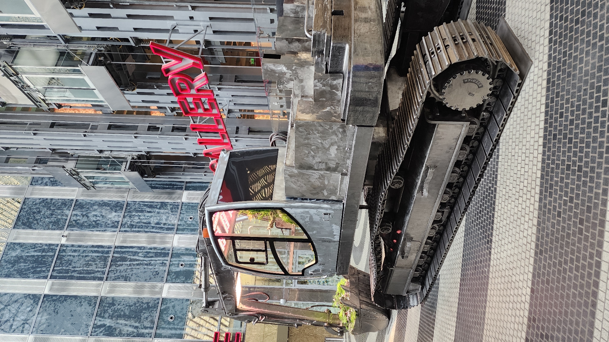 The image shows an excavator positioned on a paved surface with a gray and white brick pattern. The excavator's tracks are visible, and its cab reflects the surrounding environment. In the background, there is a modern building with a glass facade and metal framework. A large red sign with the word "Алексей" in Cyrillic is also visible, indicating the location is likely Russia. Based on the architectural style and the presence of the Cyrillic sign, it is likely that the location is in the city of Moscow, Russia. The image captures a construction scene in an urban setting.