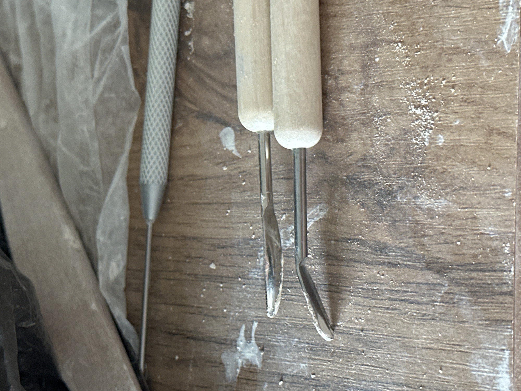 Two pottery carving tools are placed on a wooden surface. One tool has a metal handle with a textured grip and a fine, pointed metal tip. The other tool has two wooden handles, each attached to a thin metal shaft that ends in a curved, flattened metal tip. The wooden surface is textured and shows visible grain. White specks, likely clay or dust, are scattered across the surface. In the background, partially visible, are a dark, soft material and a crinkled translucent sheet. The scene appears to be an indoor workspace. There is no indication of the city of Nevinnomyssk, Russia.