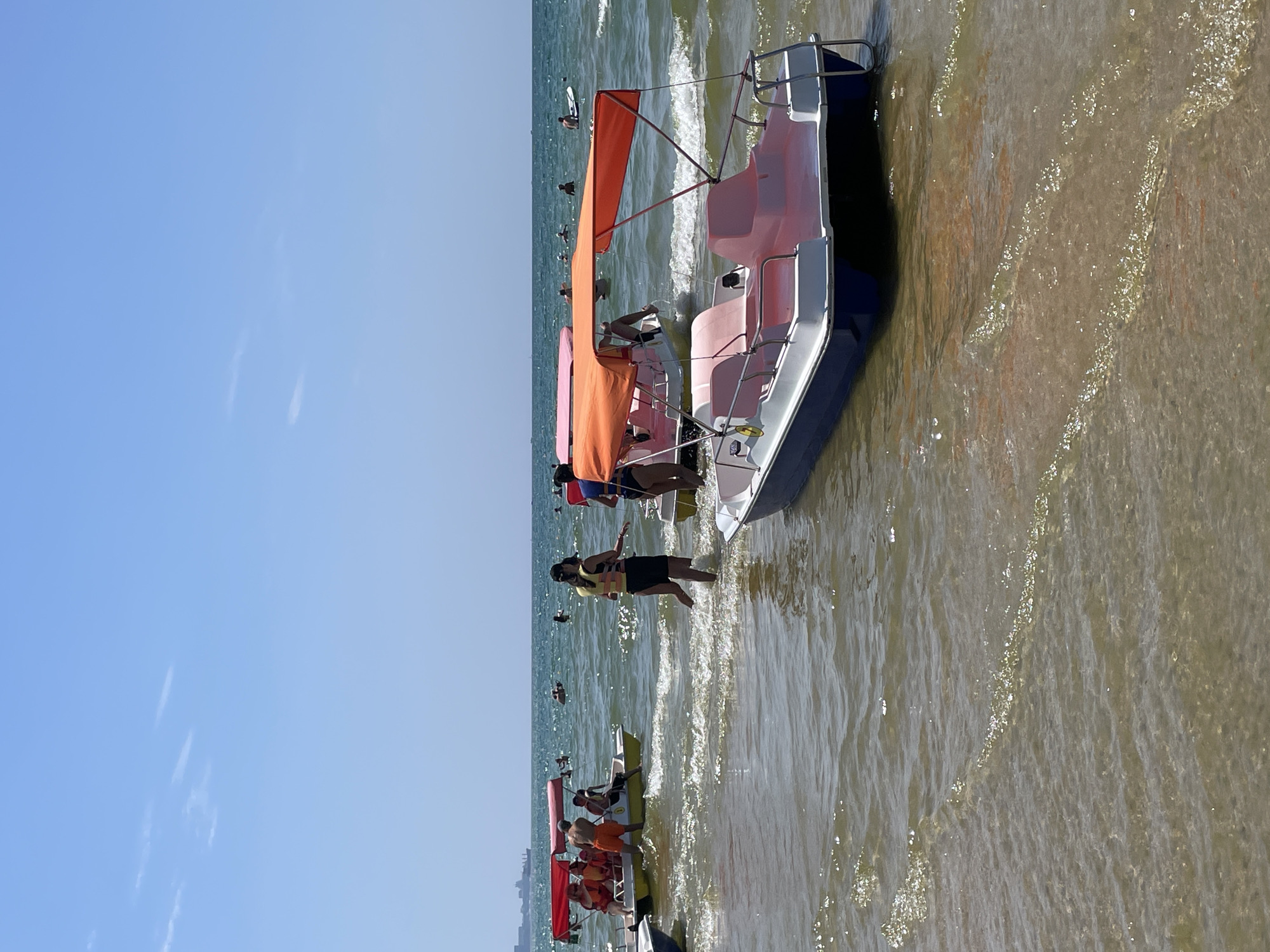 In Anapa, Russia, on a clear, sunny day, several paddle boats are positioned in the shallow, clear waters of the Black Sea near the shore. The water is calm with small waves. In the foreground, a blue and white paddle boat with pink seating and an orange canopy is partially submerged. Further out, another similar paddle boat with a pink canopy is occupied by a person. To the left, a yellow paddle boat with a red canopy is carrying multiple people, some of whom are wearing life vests. In the background, several people are swimming in the sea, and a faint outline of a city skyline is visible on the horizon. The overall scene depicts a typical summer day at a crowded beach resort.