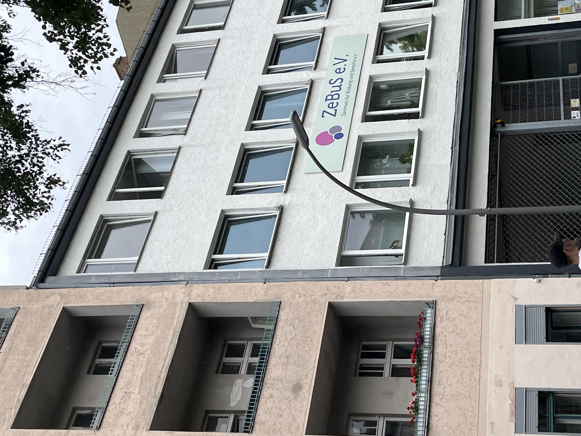 A two-story building with an adjoining apartment building is visible in Berlin, Germany. The apartment building on the left features balconies with recessed windows and flower boxes on one level. The building on the right has a white, textured facade with multiple windows. A streetlamp is positioned in front of the right building, and a sign reads "ZeBus e.V. Zentrum für Bildung und Sprache e.V.". The sky is overcast.