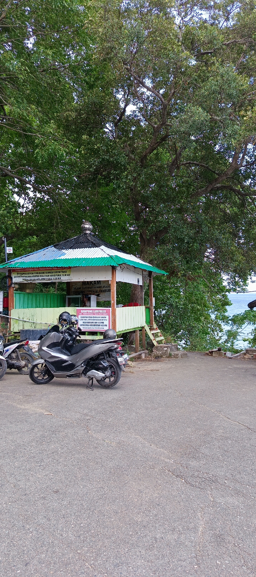 The image depicts an outdoor scene in Lampageu, Indonesia, near the sea. A small, elevated pavilion with a green and white striped roof stands prominently in the foreground. The pavilion houses a sign that reads "MAKAM SYEKH SUFIUSTRWAN ATJEH SYEKH AL-FANSUR." In front of the pavilion, two scooters are parked on an asphalt surface. The larger scooter, a dark grey automatic, is in the center, and a lighter colored scooter is partially visible to its left. A large tree with dense green foliage dominates the background, casting shadows and creating a natural canopy. Beyond the tree and pavilion, a body of water, likely the sea, is visible, with a hint of coastline. The overall atmosphere is tranquil and suggests a place of historical or religious significance.