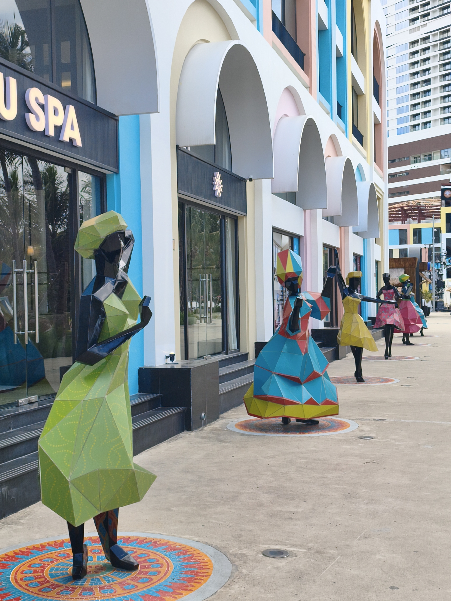 The image depicts a row of stylized sculptures of women in brightly colored, geometric outfits standing in front of a building in Nha Trang, Vietnam. The building features a modern architectural style with pastel-colored facades, arched entrances, and a large hotel or apartment building in the background. The sculptures are in a polygonal art style, with one prominent sculpture in the foreground wearing a lime green outfit. The ground is paved with concrete and decorated with circular, mosaic-like patterns beneath each sculpture. The overall scene suggests an outdoor public space or a promenade, likely a tourist attraction or shopping area.