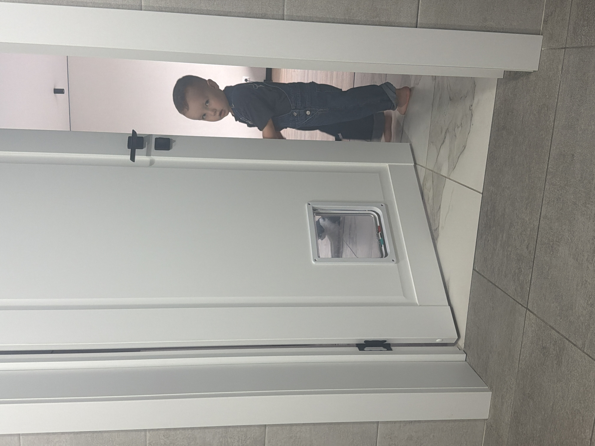 A small child, presumably a toddler, wearing dark denim overalls is crawling through a doorway. The door is white with black hardware and a small, framed pet door installed at eye-level. The background shows a bright, presumably tiled, interior space on one side of the doorway, and gray tiled walls on the other. The exact location is unknown, but it is claimed to be in Chaikovsky, Russia.