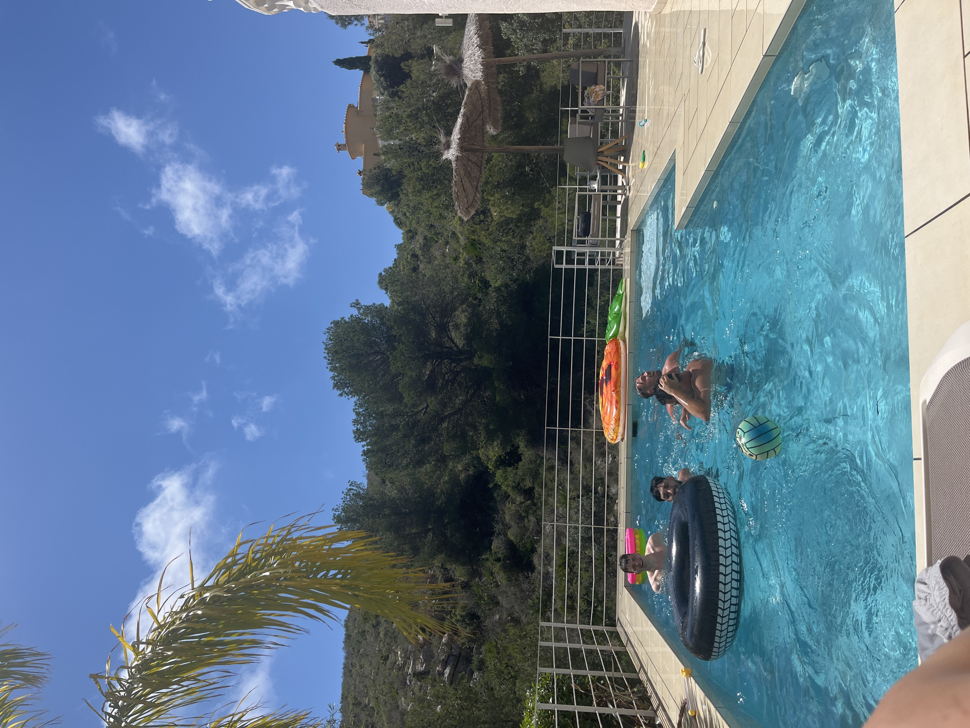 The image captures a sunny day at a swimming pool in what appears to be El Poble Nou de Benitatxell / Benitachell, Spain, suggested by the surrounding Mediterranean vegetation and architecture. The pool, filled with clear blue water, contains several people enjoying recreational activities. Three individuals are visible in the water: one man is swimming near an orange inflatable ring, another man is floating inside a black tire tube with a pink and green inflatable ring around his head, and a third person is in the distance, but not identifiable. A green and blue striped ball floats near the people in the water. Poolside furniture, including a table and chairs, are seen in the background under a straw umbrella. A metal railing separates the pool from the surrounding greenery and hillside. The sky is blue with scattered white clouds. In the foreground, a portion of a palm tree frond is visible.