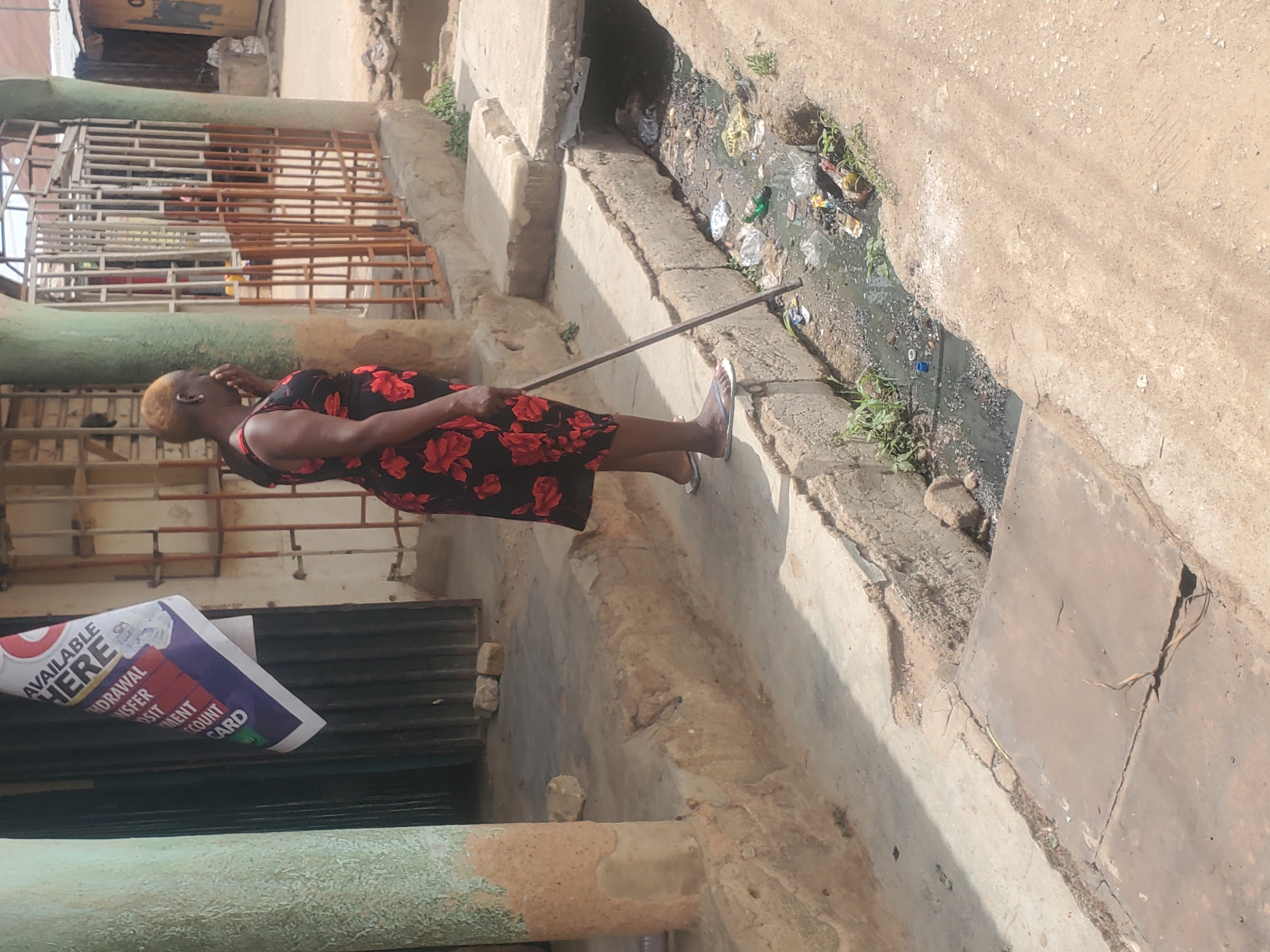 An adult woman wearing a black dress with red floral print is standing next to a concrete drainage ditch filled with stagnant water and refuse. She is using a metal rod to poke at something in the ditch. The area appears to be urban, with buildings nearby, one displaying a sign indicating available financial services. The location is Jos, Nigeria.