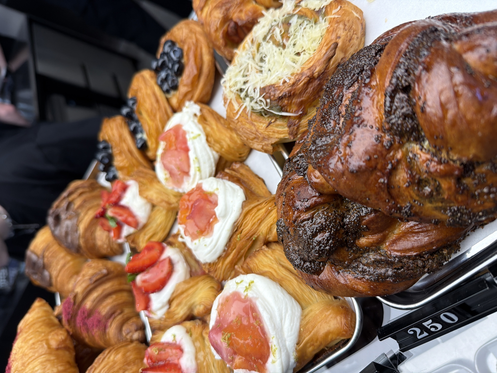 This media file shows a close-up, top-down view of a tray filled with assorted pastries, likely from a bakery or food stall. The pastries vary in type and decoration. Some appear to be croissants, one with a dusting of pink powder. Several pastries are topped with white cream and pieces of fruit, possibly strawberries or a strawberry compote, and a sprinkle of lime zest. Another pastry is topped with what looks like shredded cheese and possibly herbs. A large, dark, braided pastry covered in poppy seeds is prominently featured on the right side of the tray. The pastries are arranged on a silver tray, and part of a black price tag with the number "250" is visible at the bottom right. In the background, out of focus, are glimpses of people dressed in dark clothing and what appears to be the interior of a vehicle or stall. The lighting suggests it could be daytime, possibly indoors or outdoors in a shaded area. The overall impression is one of a delicious selection of baked goods being displayed for sale. There is no clear indication of time of day, weather, or emotions. The location context provided is Izhevsk, Russia.