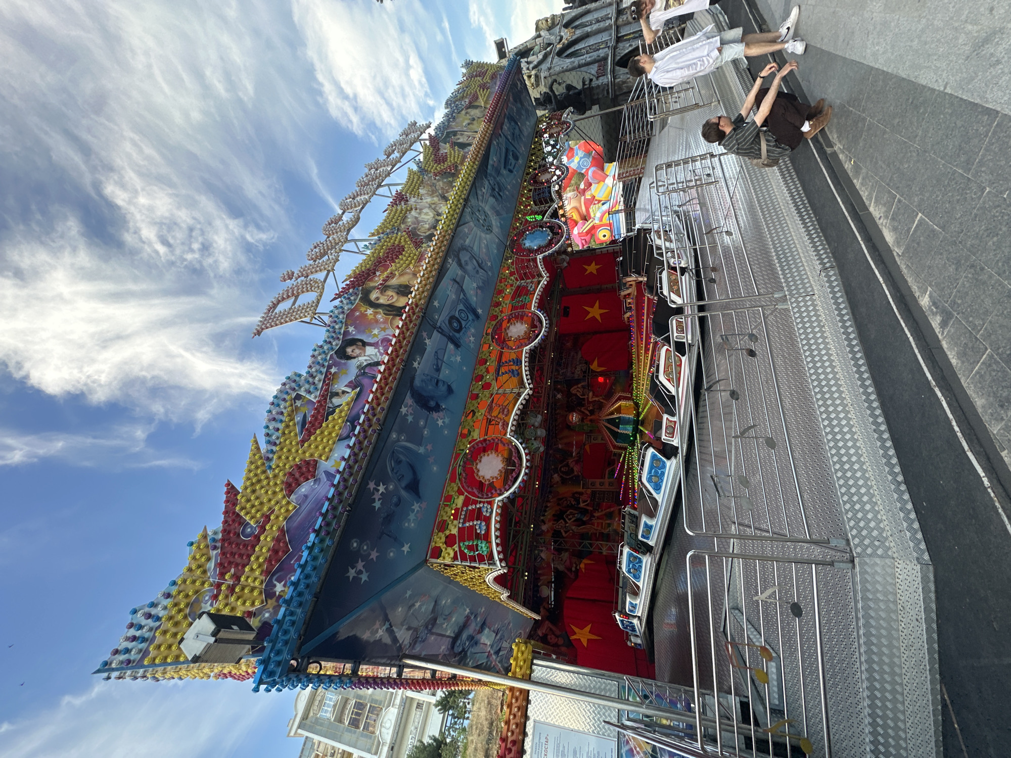 This media file depicts a vibrant amusement park ride under a bright, cloudy sky. The ride is a multi-colored structure adorned with numerous lights and decorative elements, including images of people and stars. Several people are visible, standing on the platform of the ride or nearby. One person is wearing a white shirt and shorts, another a dark striped shirt and pants, and a third a white shirt. The scene appears to be in an outdoor, possibly urban, setting, indicated by the pavement and a glimpse of buildings and trees in the background. The overall atmosphere suggests a lively and fun-filled day at an amusement park. The time of day appears to be daytime, with the sun casting light on the scene. There are no discernible emotions captured from the people in the image. Some text is visible on a sign on the ride, but it is not legible.