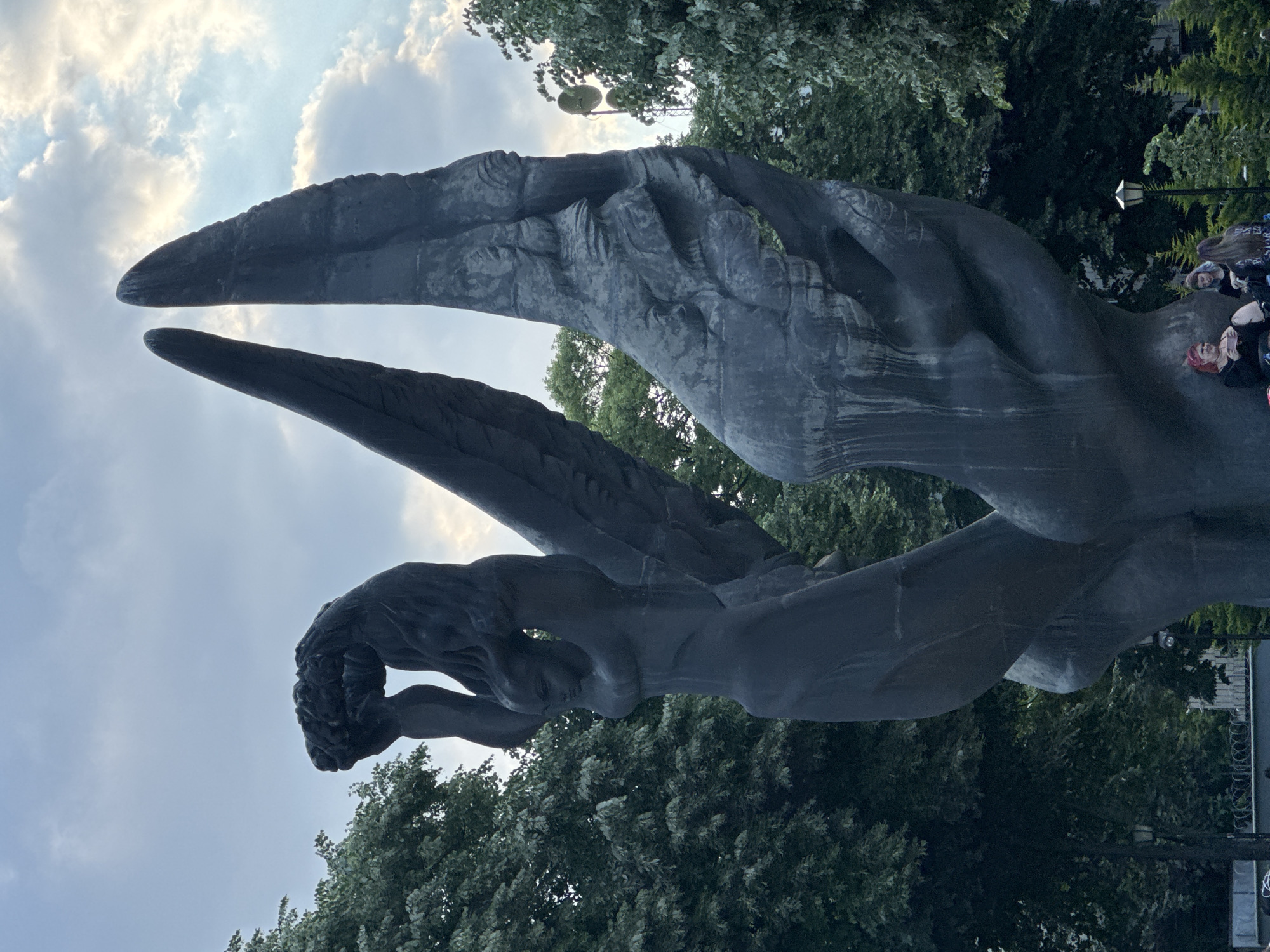 The image shows the Monument of Gratitude to the Soviet Army, also known as Alyosha, in Plovdiv, Bulgaria. The monument features a stylized female figure with outstretched arms, possibly representing a winged angel or allegory of freedom. It's constructed from a dark material, likely concrete or bronze. The sky in the background is cloudy. Trees surround the monument, giving a park-like setting. Some people are present towards the right of the image. The monument is viewed from a low angle, looking upward.