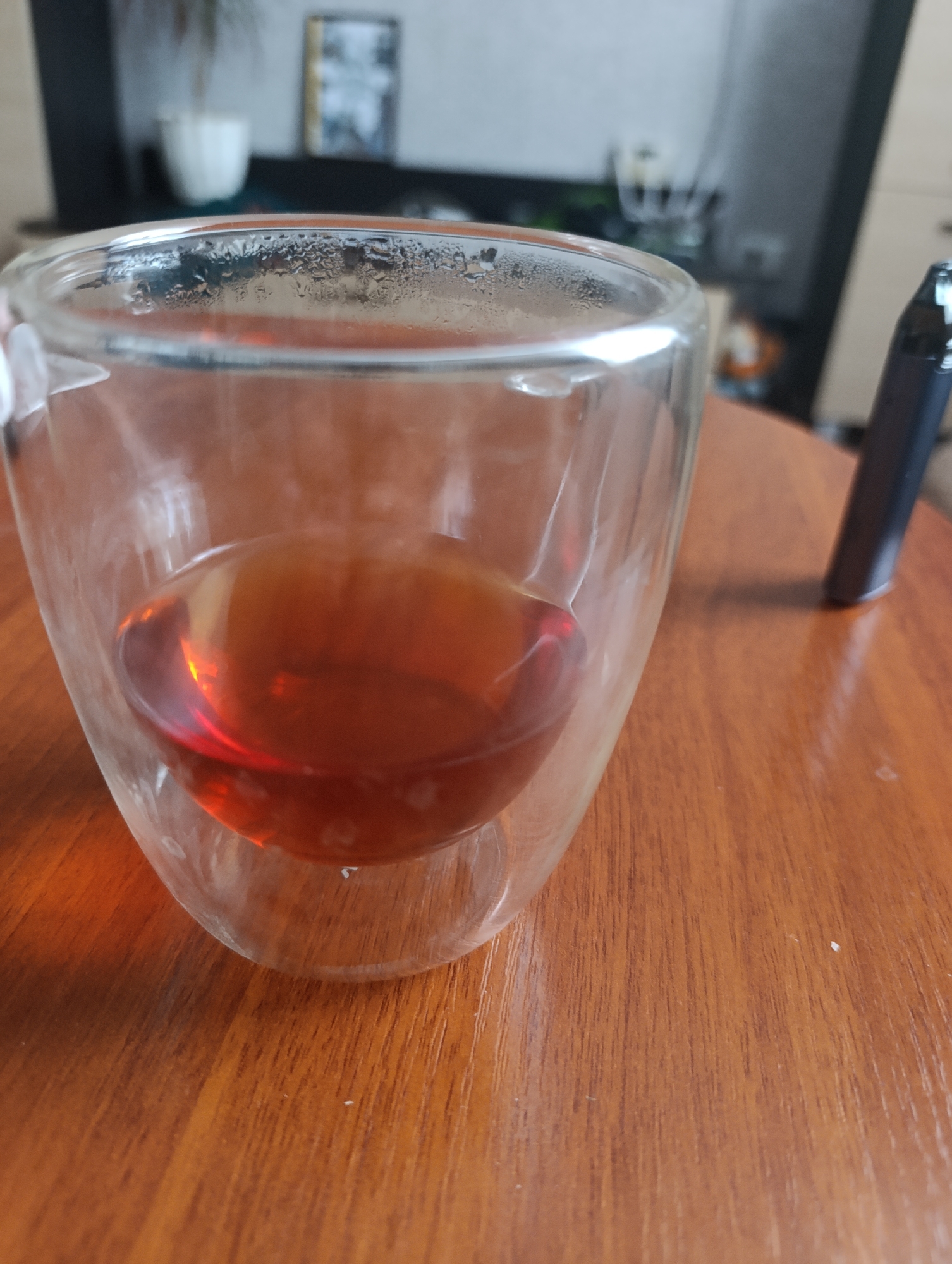 In an indoor setting, a double-walled glass containing a reddish-brown liquid, likely tea or coffee, sits on a wooden table. Condensation is visible on the exterior of the glass. To the right of the glass, a dark, slender object resembling a vape pen is partially visible. The background is blurred, showing elements of a room, including a framed picture and a white potted plant. The scene appears to be a personal space, possibly a home or office.