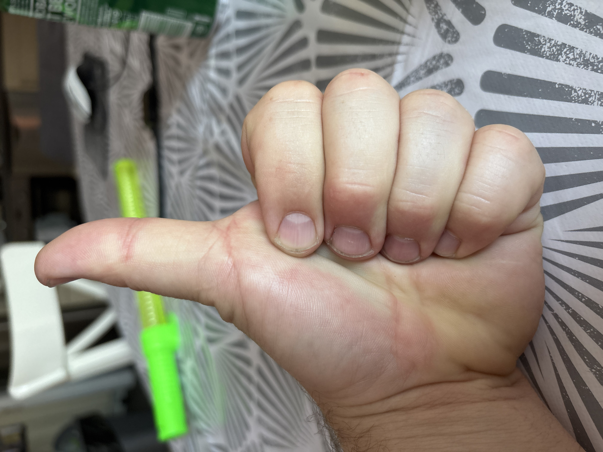 This is a close-up, eye-level photograph of a person's right hand giving a thumbs-up gesture. The hand is positioned in the foreground, obscuring most of the background. The thumb is extended upwards, and the fingers are curled inwards, forming a fist. The skin on the back of the hand and fingers appears lightly tanned and has visible creases and lines. The fingernails are short and appear clean. The background is out of focus, showing a patterned tablecloth, a white chair, and some objects that are not clearly identifiable, including a green item that may be a can or bottle and a green cylindrical object. The overall lighting suggests an indoor setting, possibly a dining area or a kitchen. The image does not contain any elements that would allow for the determination of its location in the city of Cork, Ireland.