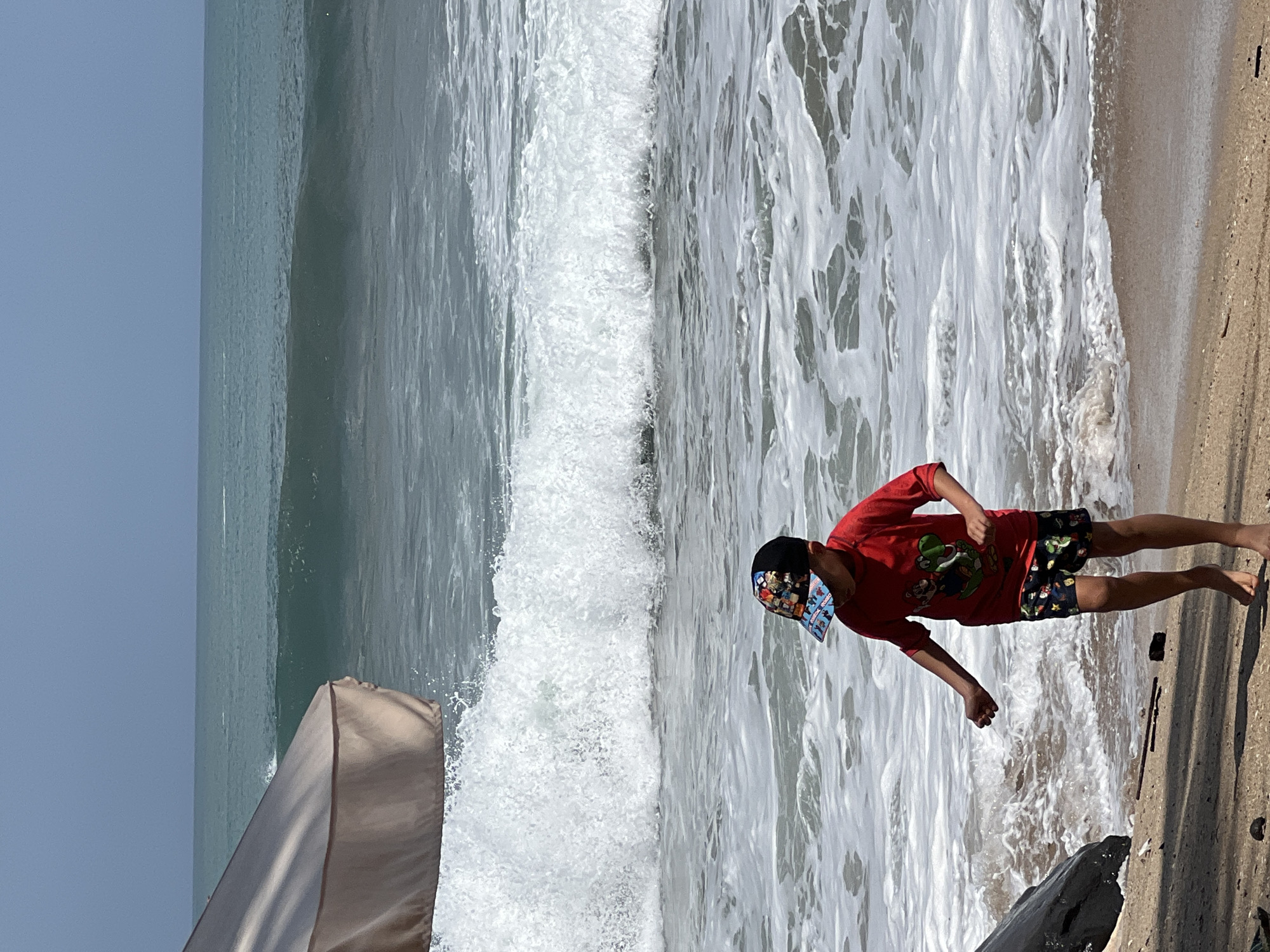 A young boy wearing a red t-shirt with a character graphic and patterned shorts is standing on a sandy beach. He has a baseball cap on his head. The boy is facing towards the ocean, with his left arm slightly outstretched. Waves are crashing behind him, creating white foam on the water's surface. In the upper left corner of the image, a portion of a large, tan umbrella is visible, casting a shadow over the scene. The ocean stretches out to the horizon, under a clear sky. The location is the beach in Kuta, Indonesia.