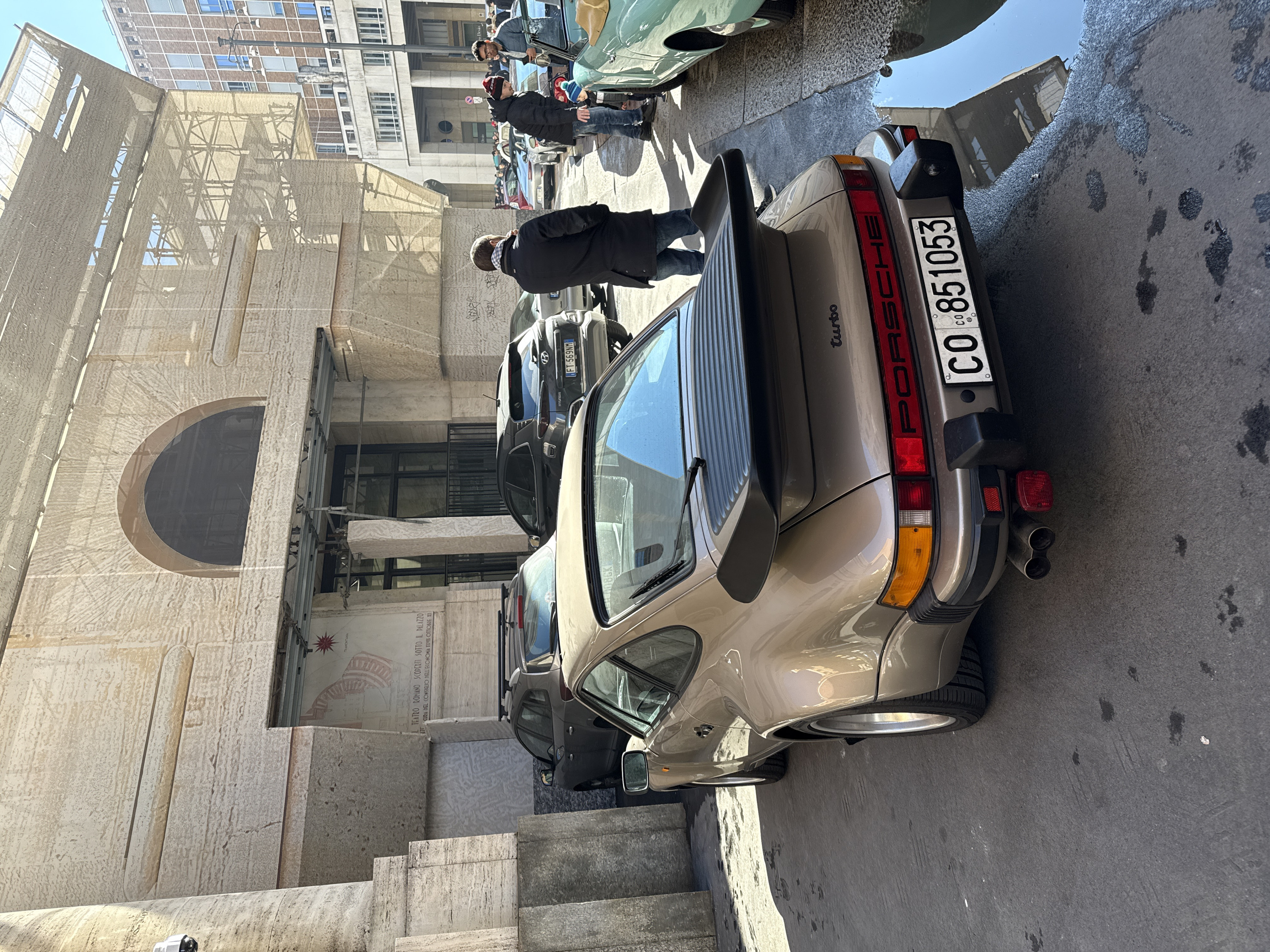 An outdoor, street-level scene in Piazza degli Affari, Milan, Italy, captures a gathering centered around vintage automobiles in front of the Palazzo della Borsa, which is partially obscured by scaffolding and tan mesh.
In the immediate foreground, facing slightly left and angled away from the viewer, is a light brown (bronze/tan) Porsche 911 Turbo, identifiable by its "whale tail" spoiler and "turbo" badging. The car features dual exhaust pipes, a black rear bumper with integrated lights, and an Italian license plate "CO 851053."
To the right of the Porsche, the front passenger side of a light green vintage convertible is partially visible. Behind these vehicles, a line of other cars is parked along the street, including a dark gray Hyundai Kona SUV with license plate "FT 569NM" and another dark gray SUV with roof rails.
Several individuals are present. A man in a dark jacket stands with his back to the viewer, to the right of the Porsche. Further down the street, near the green car, a man in a dark jacket and a red knit hat leans down towards a child wearing a blue and white striped knit hat. Beside them, another man in a light blue jacket and glasses observes the scene, holding a small object. Additional people are visible in the distant background.
The ground is paved with asphalt, showing various dark stains, possibly from oil or water, and a noticeable puddle reflecting the sky and buildings in the lower right quadrant. The large, light-colored stone (Travertine) building in the background, the Palazzo della Borsa, features prominent columns and an arched entry point. A security camera is mounted on a column on the far left. A poster with Italian text referencing "IL TEATRO ROMANO SCOPERTO SOTTO IL PALAZZO" is visible within an arched opening on the building. Across the street on the right, another multi-story urban building with a grid pattern of windows is visible under a clear sky. Sunlight casts distinct shadows across the street.