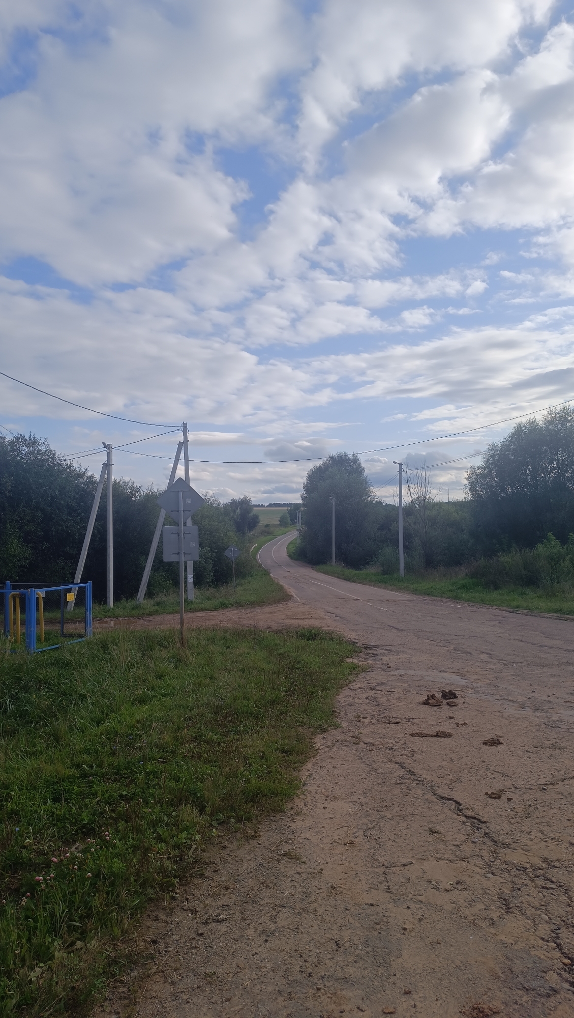 The image depicts a rural road in the city of Karsha, Russia, on a partly cloudy day. A paved road curves to the left, disappearing into a landscape of trees and fields. The road surface shows signs of wear and tear, with cracks and patches visible. To the left of the road, there is a grassy verge with a yellow and blue metal barrier. Several utility poles with wires are positioned along the roadside, and a road sign is mounted on one of them. The sky is filled with white and grey clouds against a blue background. The overall impression is one of a quiet, unpopulated rural setting.
