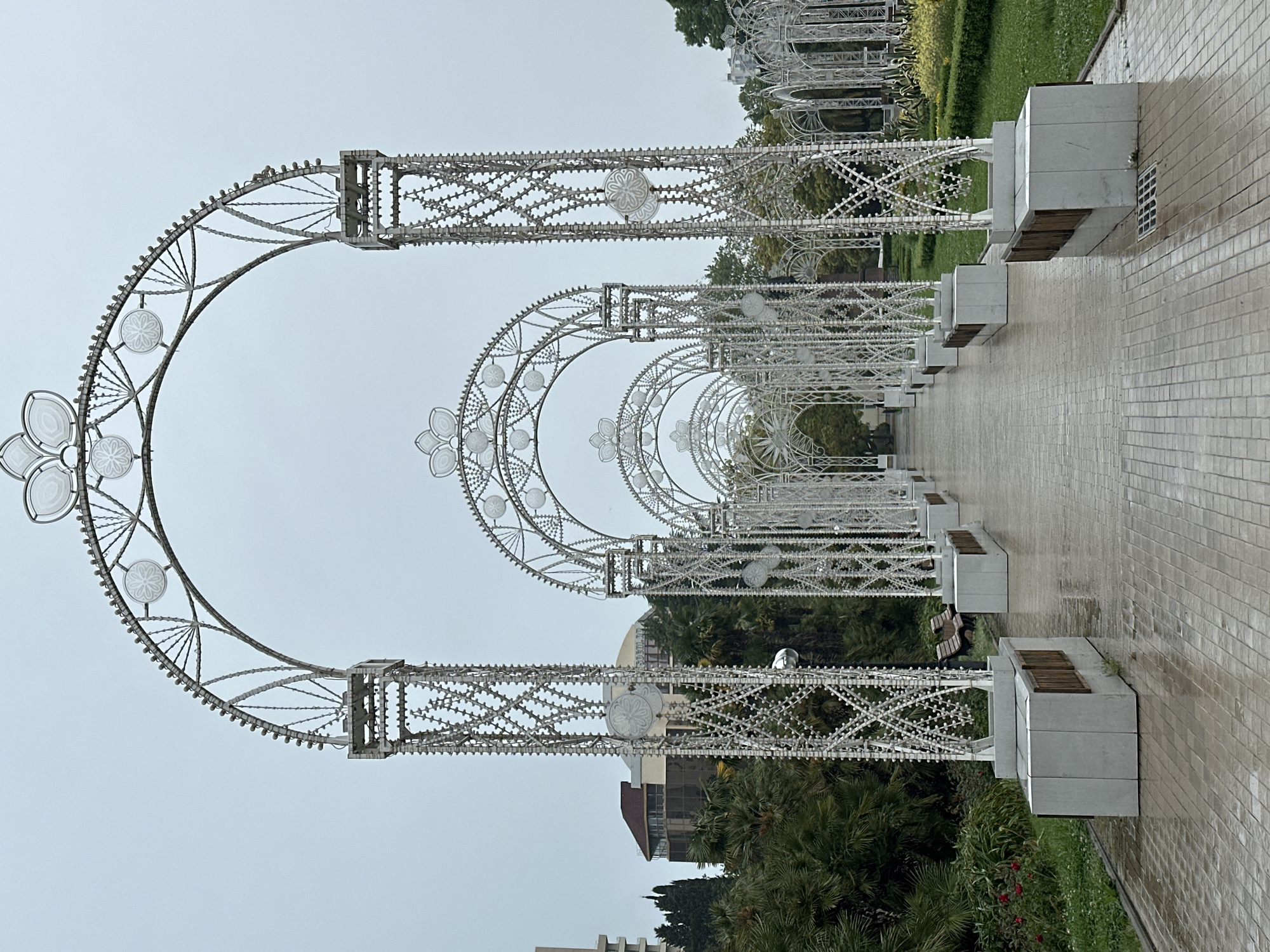 The photograph shows a colonnade of illuminated metal arches located in Sochi, Russia. The arches are white and decorated with floral patterns. The colonnade forms a pedestrian pathway with benches along the sides. The ground is paved with light-colored bricks, appearing wet, suggesting recent rain. Lush green trees and vegetation are visible behind the arches, indicating a park setting. The sky is overcast, creating a diffuse light. A building is visible in the background.