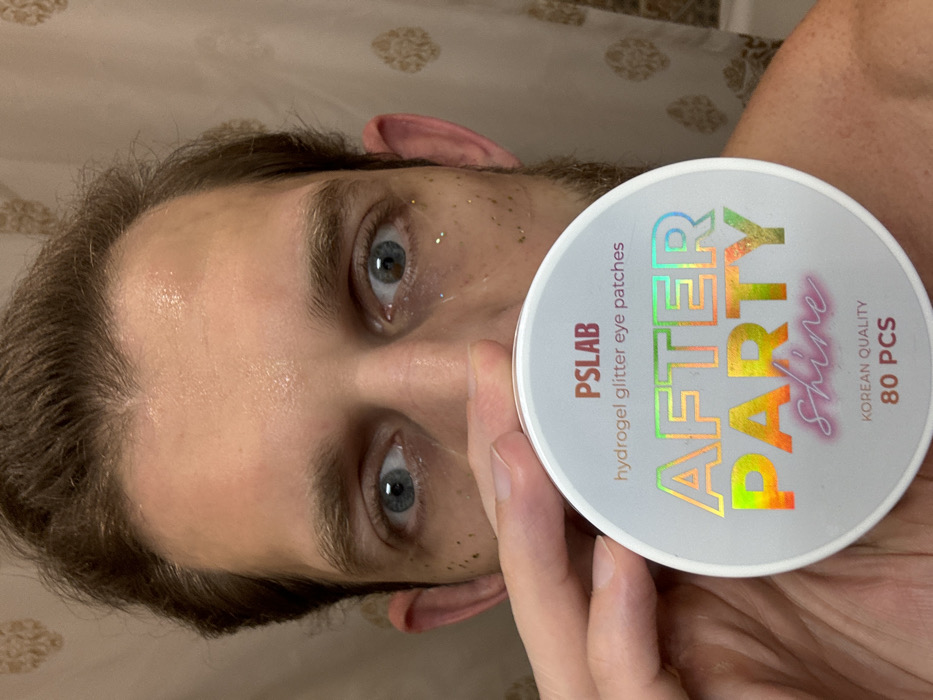 A person is holding a container of "PSLAB hydrogel glitter eye patches AFTER PARTY Shine" in front of their face. The container is white with iridescent lettering. The person's eyes are visible, and there are small pieces of glitter under one eye. The person has light brown hair. The container says "Korean Quality" and "80 PCS".