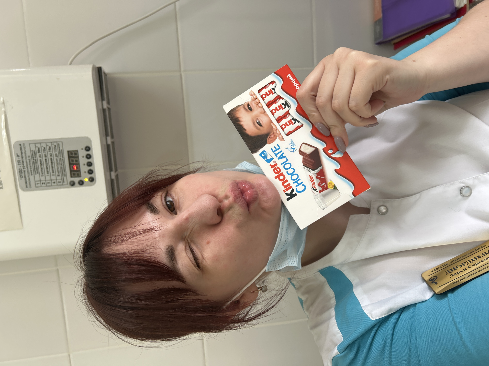 A woman in a white and blue medical uniform is holding a Kinder Chocolate bar in a room with white tiled walls. She is wearing a light blue face mask and making a pouting face with one eye closed. A name tag on her uniform reads "ДИОРДИЕВА Дарья Сергеев", identifying her as a nurse at "ГБУЗ ПК «Чайковская ЦГБ». A piece of equipment with a digital display is visible on the wall behind her. The location mentioned in the OCR is Чайковская, Russia.
