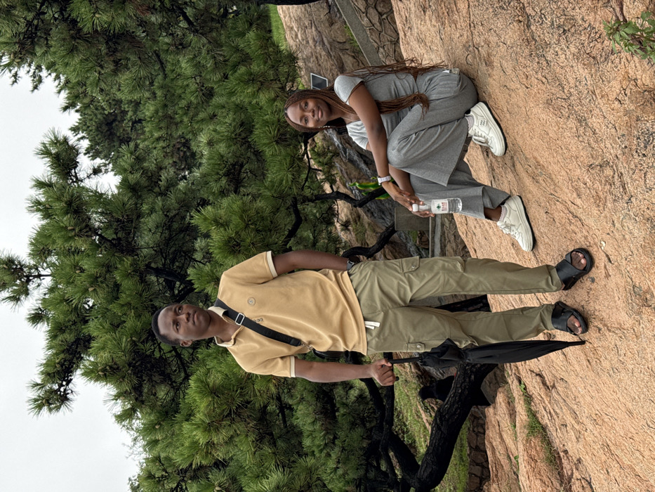 This image depicts an outdoor scene in Shinan District, China, featuring two individuals amidst a rocky landscape with pine trees under an overcast sky. On the left, a male subject stands, positioned in a three-quarter profile facing right. He wears a tan polo shirt with a lighter collar, khaki cargo pants with a white drawstring, and black cross-strap sandals. A black cross-body bag is worn across his chest. His left hand holds a closed black umbrella, while his right is in his pocket. On the right, a female subject sits on a large reddish-brown rock, facing slightly left with her gaze directed towards the viewer. She is dressed in a grey short-sleeved top and matching wide-leg pants, paired with white sneakers. A silver-colored watch is visible on her left wrist. She holds a clear plastic water bottle with a white label, bearing Chinese characters, in her right hand. The immediate ground consists of varied reddish-brown rock formations, sloping gently upward. In the mid-ground, behind the female subject, a small, dark green decorative element is partially visible. The background is densely populated with mature pine trees, displaying dark green needles and visible dark, gnarled branches.