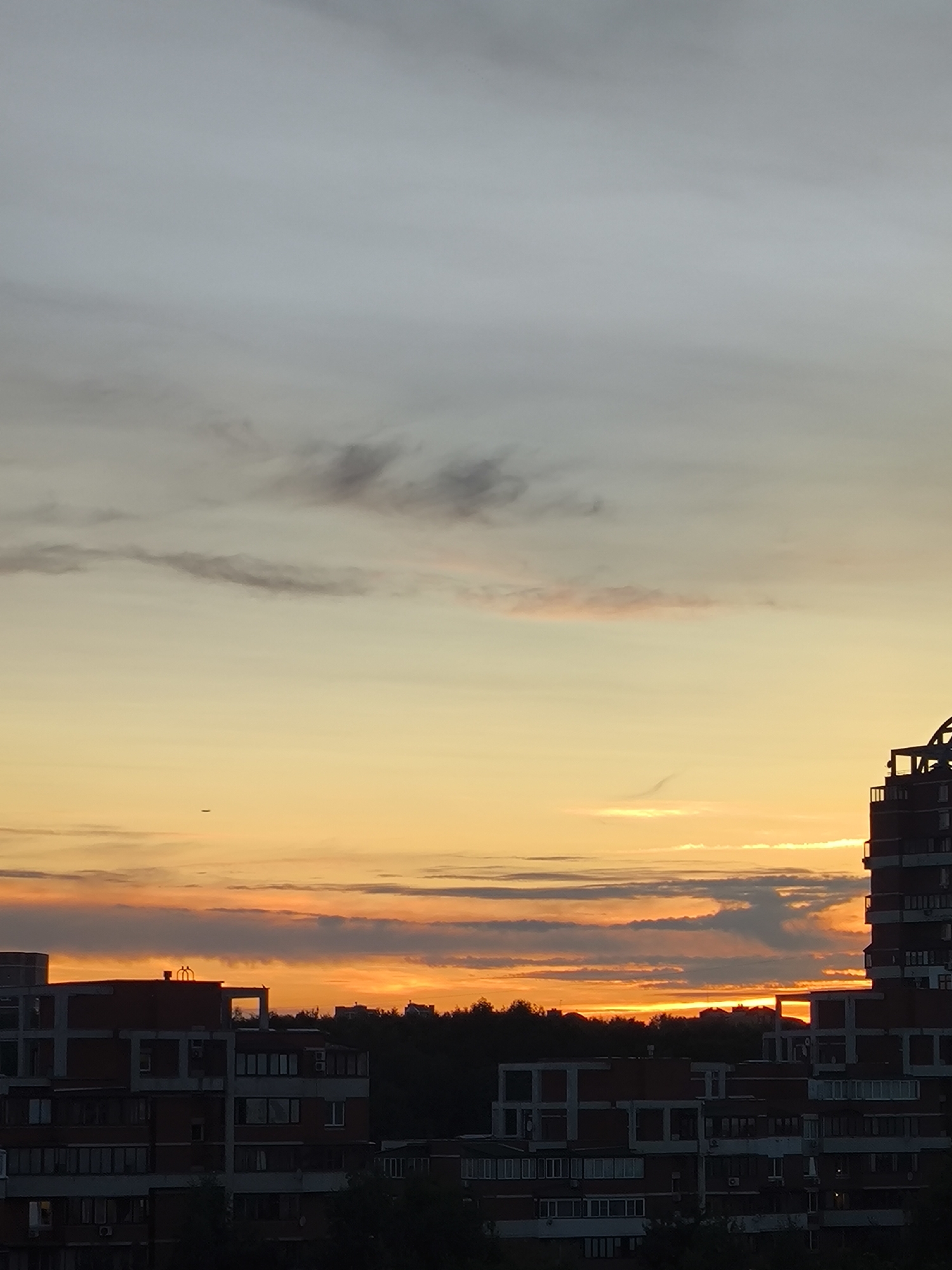 The image shows a sunset over a city. The sky is filled with shades of orange, yellow, and gray. The sun is low on the horizon, casting a warm glow over the scene. In the foreground, there are several buildings, some of which appear to be under construction. The buildings are made of brick and have a modern design. Trees are visible between the buildings, suggesting a mix of urban and natural landscapes. A small airplane can be seen flying in the distance. The overall atmosphere is peaceful and serene, capturing the beauty of a city at dusk.