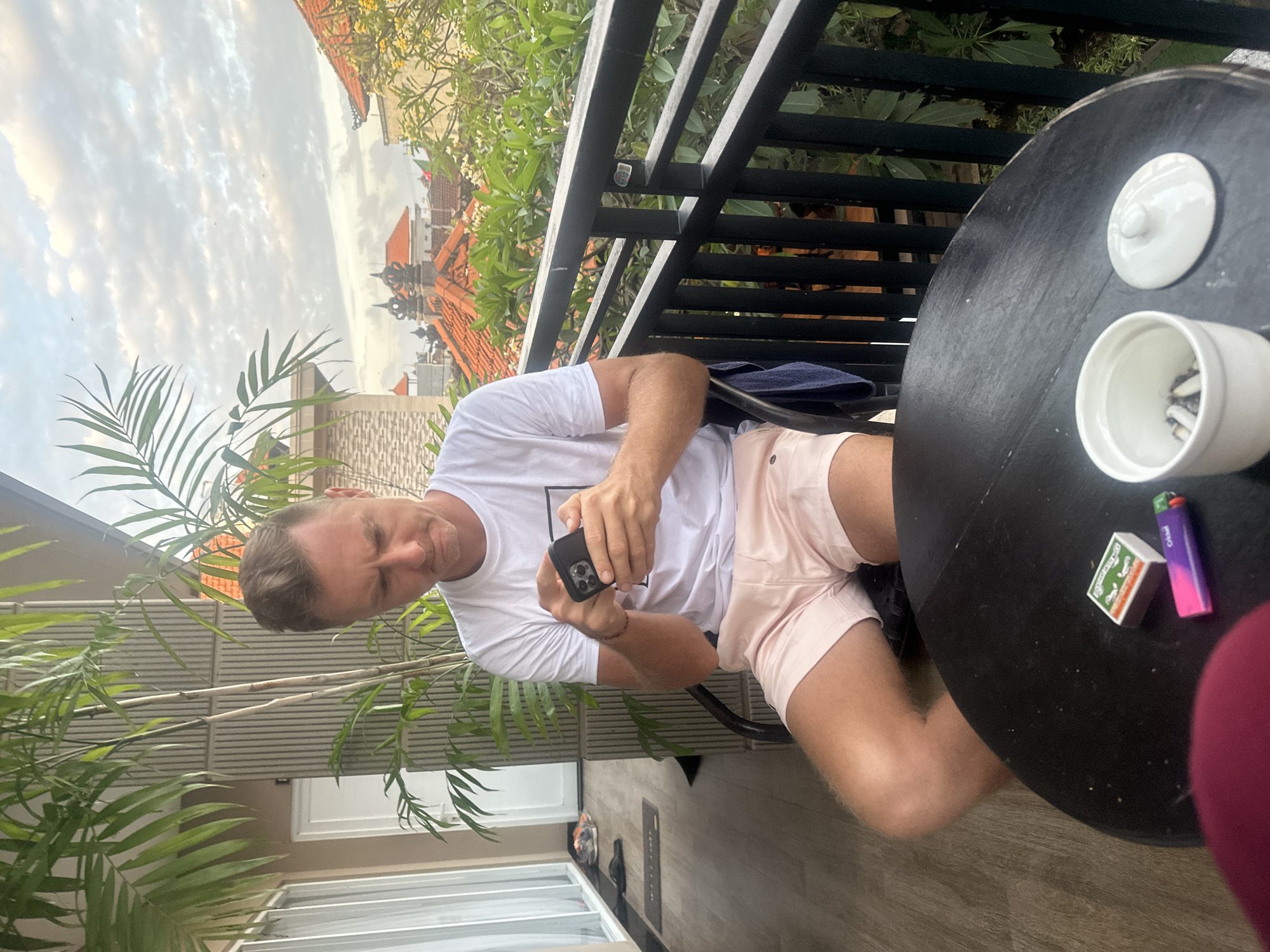 A man with blond hair, wearing a white t-shirt and pink shorts, is sitting at a black circular table on a balcony. He is looking down at his smartphone, which he is holding in both hands. On the table, there is a small white ashtray with cigarette butts, a matchbox, and a lighter. A dark blue towel is draped over the back of his chair. Behind him, palm fronds are visible, and further in the background, red-tiled roofs of buildings, characteristic of Kuta, Indonesia, are seen under a cloudy sky.