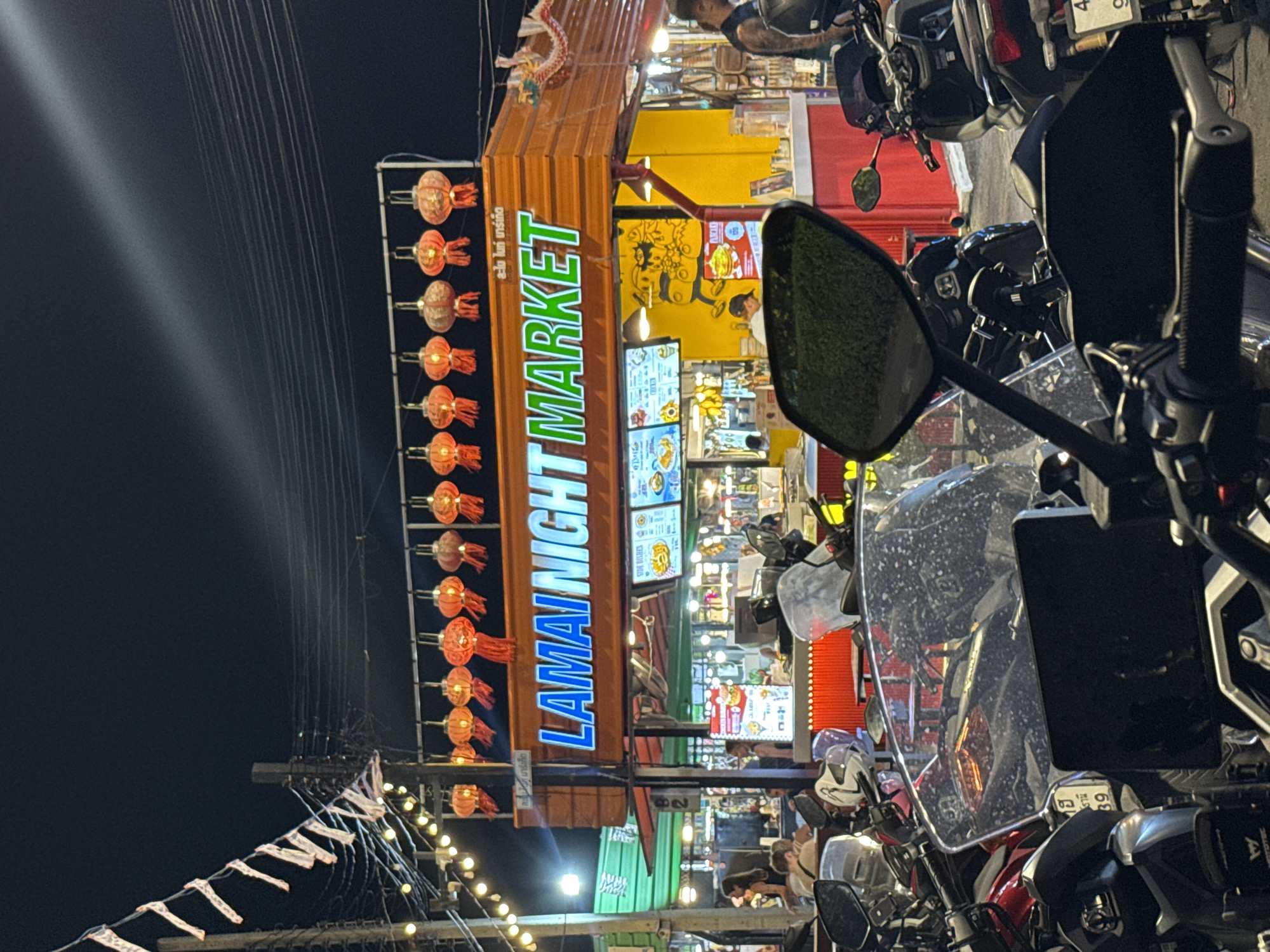 This image captures a vibrant night scene at the Lamai Night Market in Baan Lamai, Thailand. The setting is lively and illuminated by numerous artificial lights against a dark night sky.
In the center, a prominent vertical blue and green neon sign spells out "LAMAI NIGHT MARKET," with Thai script "ละใน ไนท์ มาร์เก็ต" visible above it. Below this sign, brightly lit market stalls display various goods and feature digital menu screens. Several people can be seen interacting with the stalls, browsing, or working, contributing to a bustling atmosphere.
To the left of the main market sign, a vertical arrangement of traditional red-orange lanterns glows warmly, adding a festive touch. Further left and extending across the upper portion of the frame, numerous utility wires or power lines crisscross the dark sky. A string of small, warm-toned fairy lights also runs horizontally across the lower-left, enhancing the market's ambiance.
In the foreground, a row of motorcycles is parked, with handlebars, mirrors, and windshields clearly visible, suggesting the perspective of someone either parking or leaving. One motorcycle features a phone mount on its handlebars.
Notable visible text includes "MOTOTEC" on a motorcycle accessory, "SIDE DISHES" on a menu board, and numbers "39," "82," and "4" on various signs or plates. The overall impression is one of a bustling, energetic market after dark.