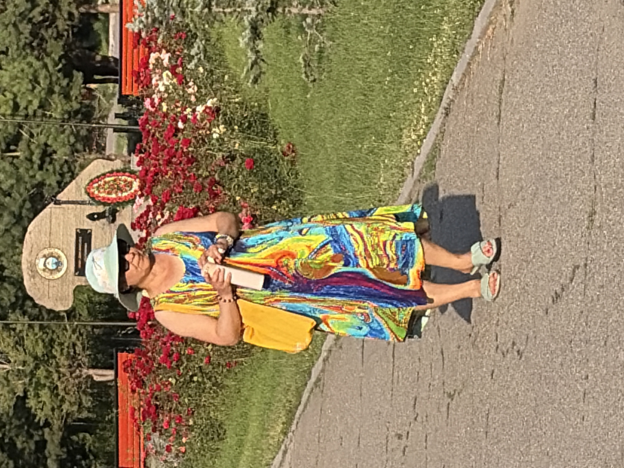 In a park on a sunny day, a woman walks on a paved path beside a grassy area and a flower bed filled with red and white roses. She wears a light blue sun hat, sunglasses, a colorful sleeveless dress, and light blue sandals. A yellow bag is slung over her left shoulder, and she holds a white water bottle in her right hand. In the background, a stone monument with a circular emblem and a wreath is visible, along with park benches and trees.