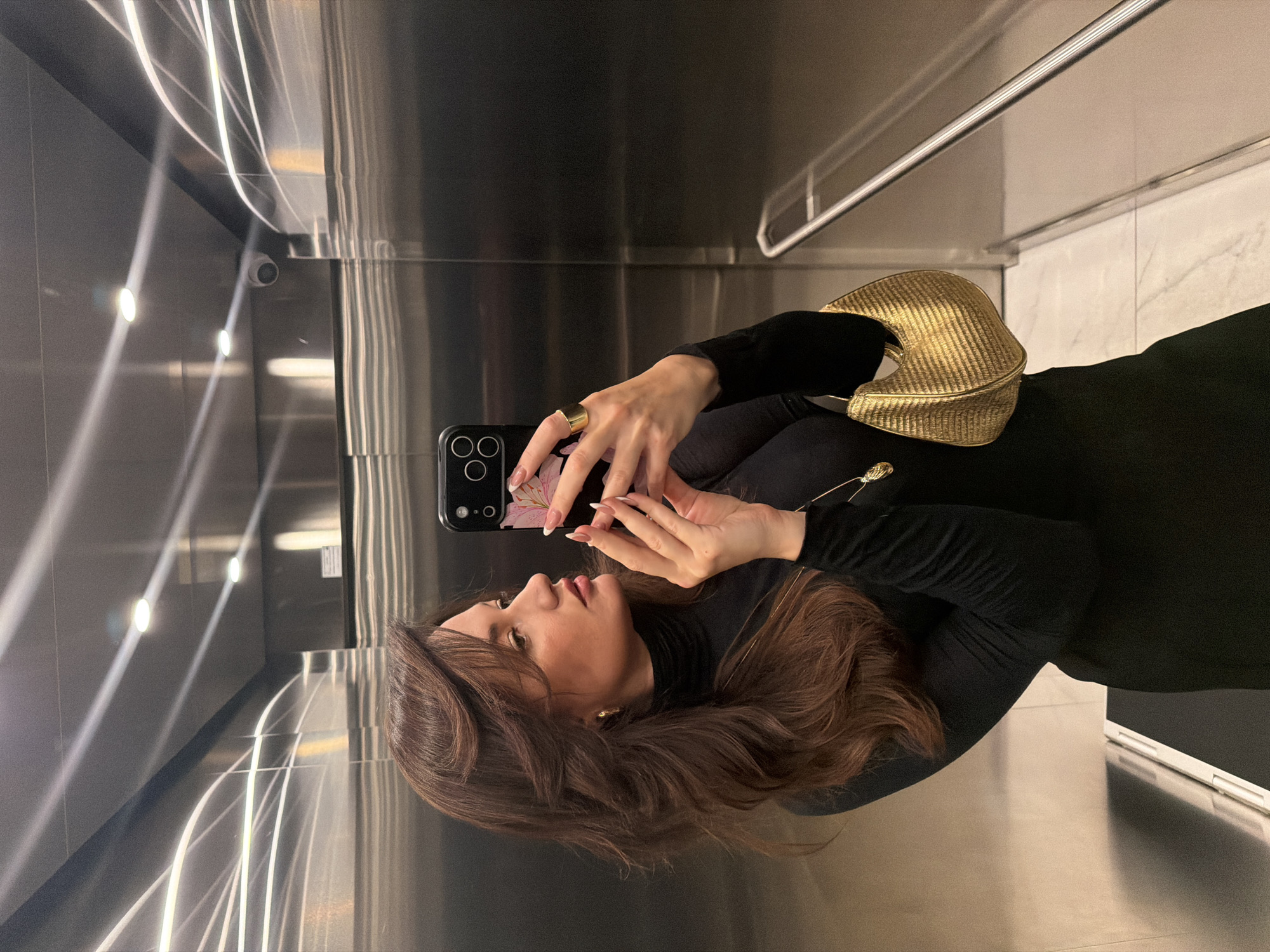 A person with brown hair and wearing a black turtleneck and dress is taking a selfie in what appears to be an elevator. The person is holding a black smartphone with a flower design on the case. A gold purse is slung over the person's shoulder. The interior of the elevator is stainless steel with vertical light fixtures, and a security camera is visible. The location is likely Belgrade, Serbia; however, this cannot be determined from the given image alone.