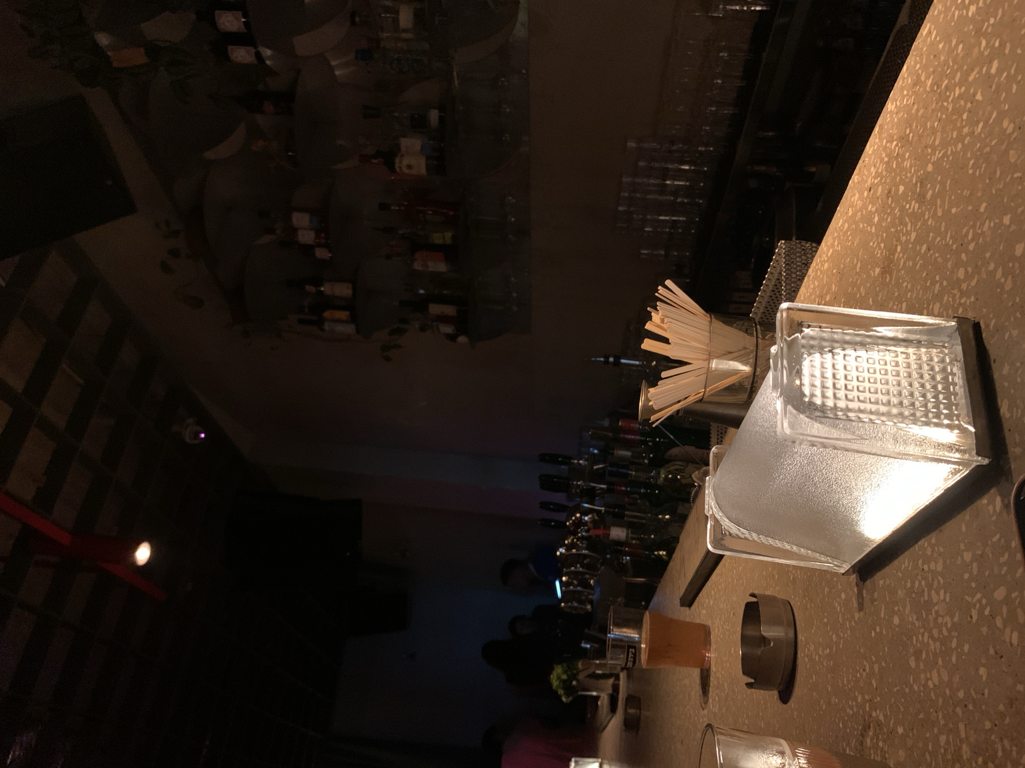 This is a photograph taken inside a dimly lit bar. In the foreground, a portion of a speckled countertop is visible, with a clear glass of amber liquid and a metal ashtray on it. To the right, a glass prism-like object refracts light. In the mid-ground, a bar with various bottles of liquor is stocked on shelves behind a row of taps. Several people are blurred in the background, suggesting activity and a social atmosphere. The overall lighting is subdued, with warm tones from the bar lights and some cooler accent lighting. The scene is consistent with a night-time setting in a bar, possibly in Tel-Aviv, Israel, given the context provided.