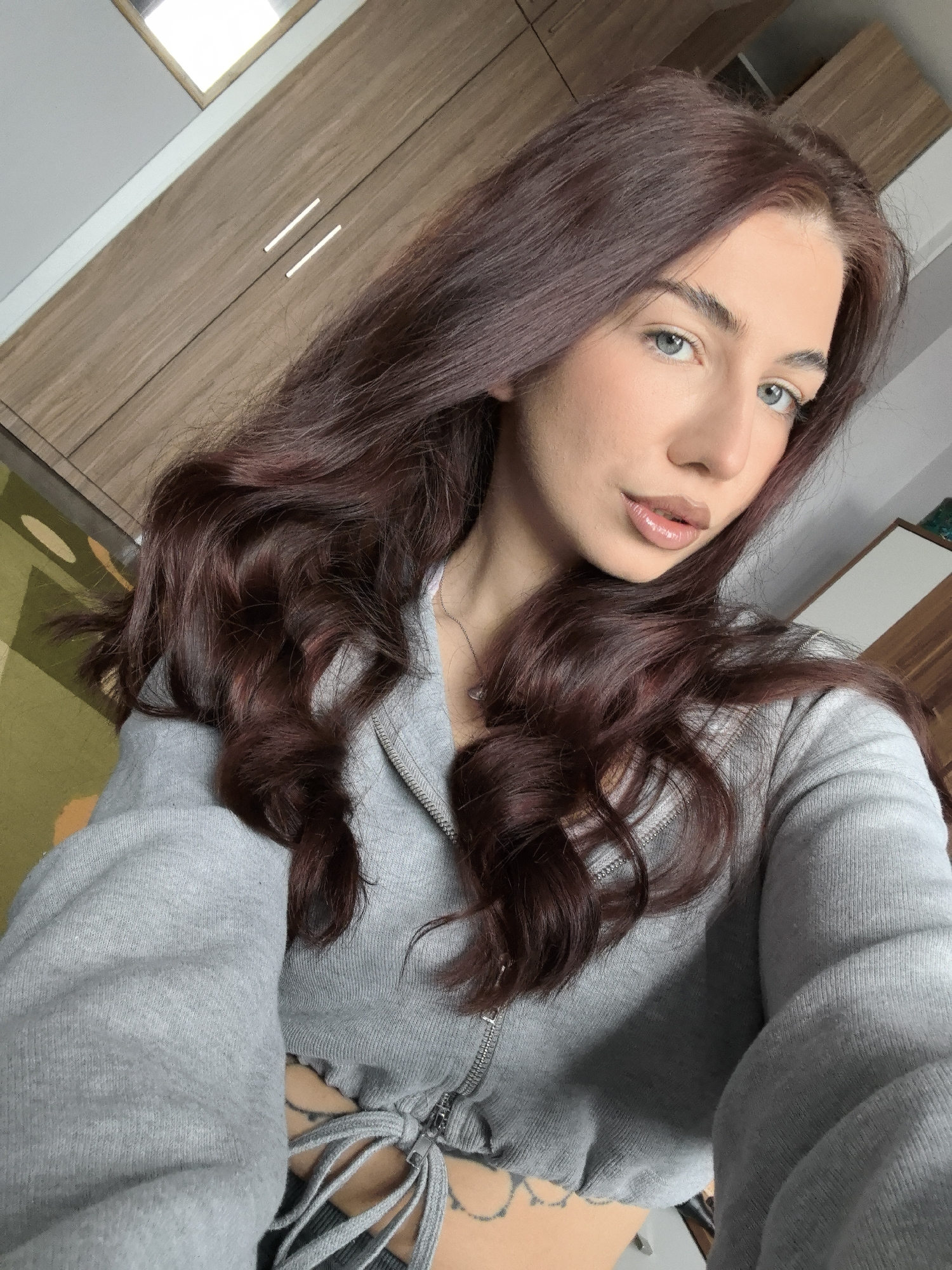 A young woman is facing the camera in a selfie. She has long, dark red-brown hair styled in loose waves, light skin, and light blue eyes. Her lips are full and appear to have a shiny lip gloss applied. She is wearing a gray zip-up hooded sweatshirt that is tied at the waist, revealing a small portion of her midriff and some tattoos on her lower abdomen.
The background includes a wooden cabinet or wardrobe, a section of a colorful patterned rug, and part of a white wall. The location is likely inside a residential building. There is no information available to determine if this is Stara Zagora, Bulgaria.