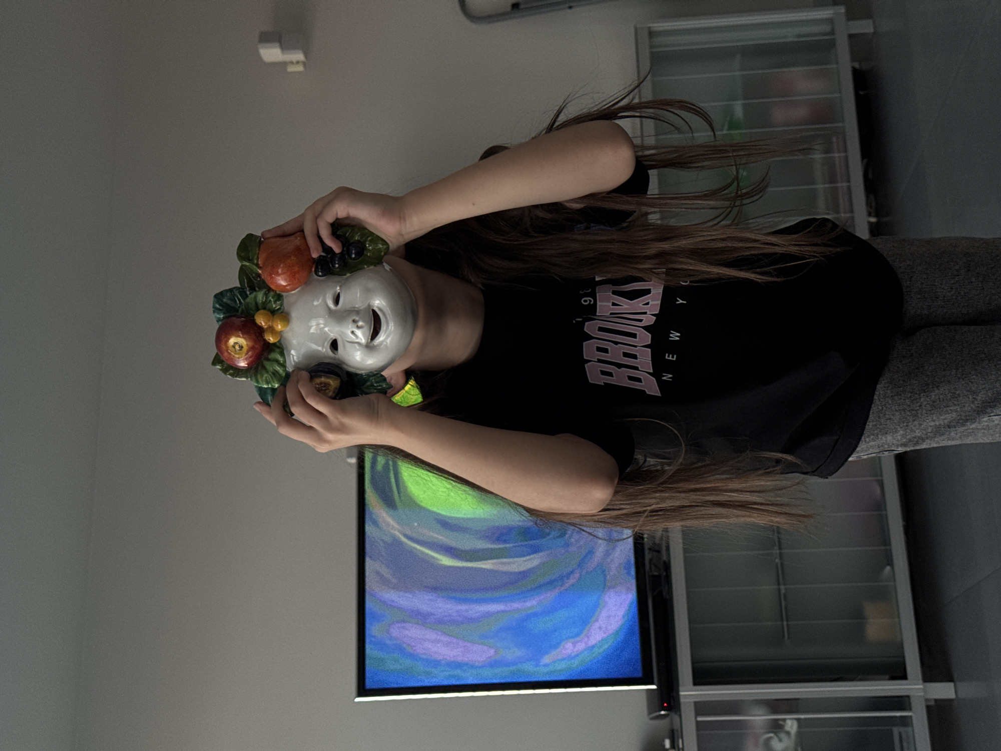 A person wearing a "BROOKLYN NEW YORK" t-shirt holds a white, fruit-adorned mask in front of their face. Behind the person is a television displaying an abstract, swirling pattern of blues, purples, and greens. To the right, a gray shelving unit is visible. The location is indoors.