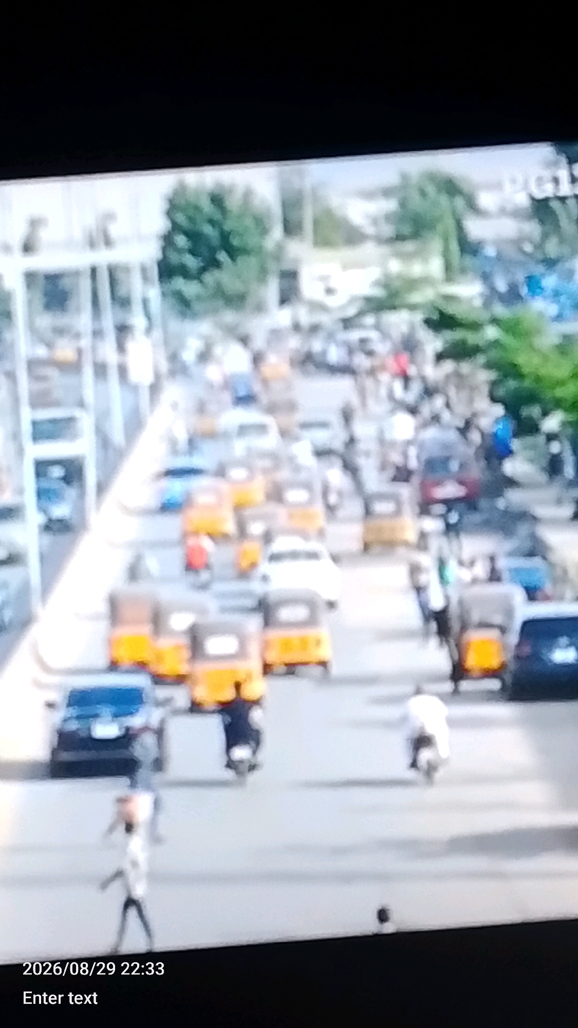 The image depicts a busy street scene in Lokoja, Nigeria, likely captured by a surveillance camera, indicated by the timestamp "2026/08/29 22:33" and the text "Enter text" at the bottom. The foreground features several yellow auto-rickshaws, commonly known as Keke Napeps, and a dark-colored car. A person in a light-colored shirt and dark pants is walking on the street, while another person on a motorcycle is visible in the mid-ground. In the background, more vehicles, including auto-rickshaws and cars, are on the road, suggesting a traffic situation. Trees are visible along the sides of the street, and some structures, possibly buildings or overhead infrastructure, are present in the distance. The overall impression is of a moderately congested urban environment.
