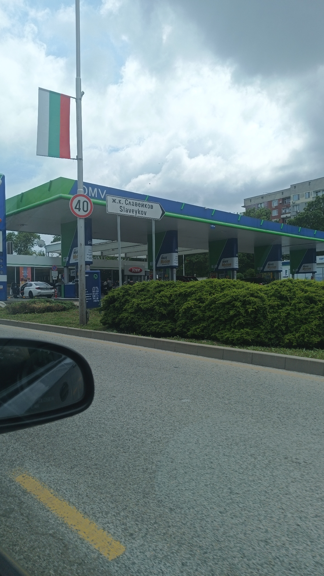 Here is a detailed description of the image:
**Location:** The image shows a scene at an OMV gas station in the city of Burgas, Bulgaria. The presence of the Bulgarian flag and the Cyrillic text on the street sign (Ж.к. Славейков - Slaveykov) confirm this location.
**Scene Details:**
* **Gas Station:** An OMV gas station with a blue and green roof is visible. Multiple gas pumps are under the station's canopy. The brand VIVA is visible near the convenience store of the station. There are "Maxx Motion" signs present on the pumps.
* **Road and Signage:** A street sign indicates "Slaveykov" in both Cyrillic and Latin alphabets, pointing to the area or direction of the Slaveykov neighborhood. A speed limit sign "40" is also visible.
* **Vegetation:** A neatly trimmed hedge runs along the edge of the sidewalk.
* **Traffic and Vehicles:** Part of a car, including the side mirror, is visible in the foreground suggesting the photograph was taken from within a vehicle. A white car is visible at one of the gas pumps.
* **People:** Some people are near the convenience store area of the station.
* **Sky:** The sky is overcast, with a mix of white and gray clouds.
* **Buildings:** In the background, an apartment building is visible.
**Overall Impression:** The photo captures a typical scene at a gas station in a residential area in Burgas, Bulgaria. The overcast weather adds to the overall ordinary atmosphere.