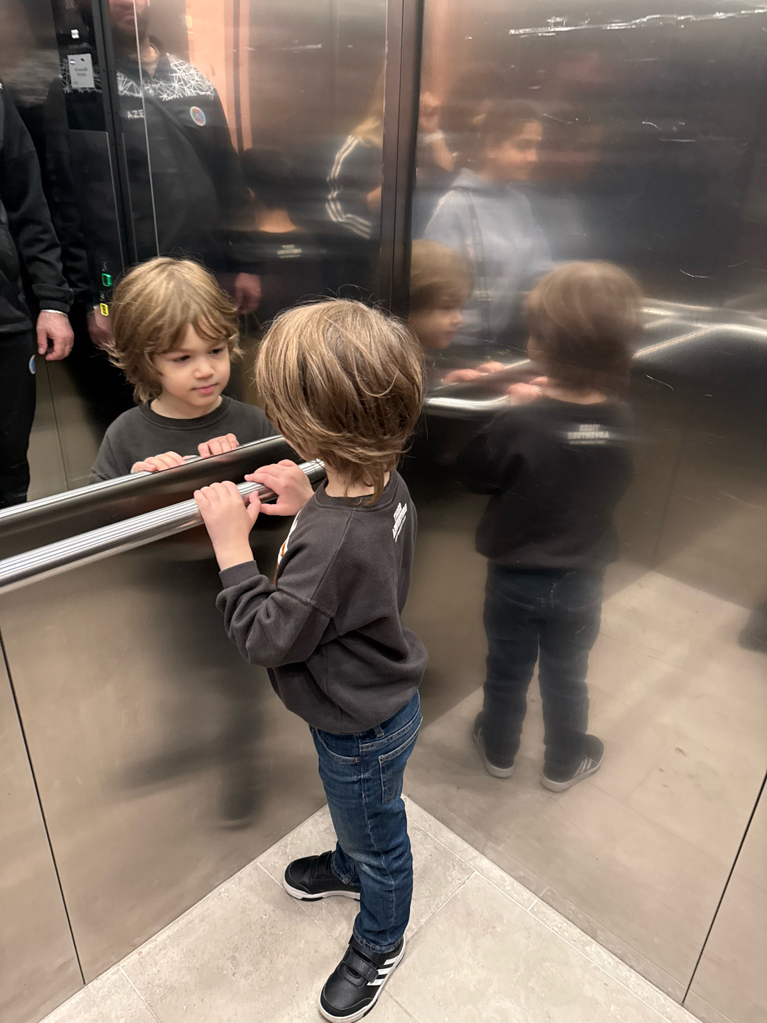 In an elevator with reflective metal walls, a young boy with shaggy brown hair stands facing a mirror, his hands resting on the horizontal metal bar. He wears a dark gray crewneck sweatshirt and blue jeans, with black and white athletic shoes. His reflection is visible, as are reflections of other individuals, one of whom is a man wearing a black jacket with a white geometric pattern and a badge, possibly a uniform. Another reflection shows a person in a light blue hooded top. The elevator appears to be in a modern building. The flooring is a light-colored tile. The scene is indoors, and based on the presence of the specific badges visible on the jacket of the man, it is likely that this elevator is located at the Tallinn Central Terminal in Tallinn, Estonia.
