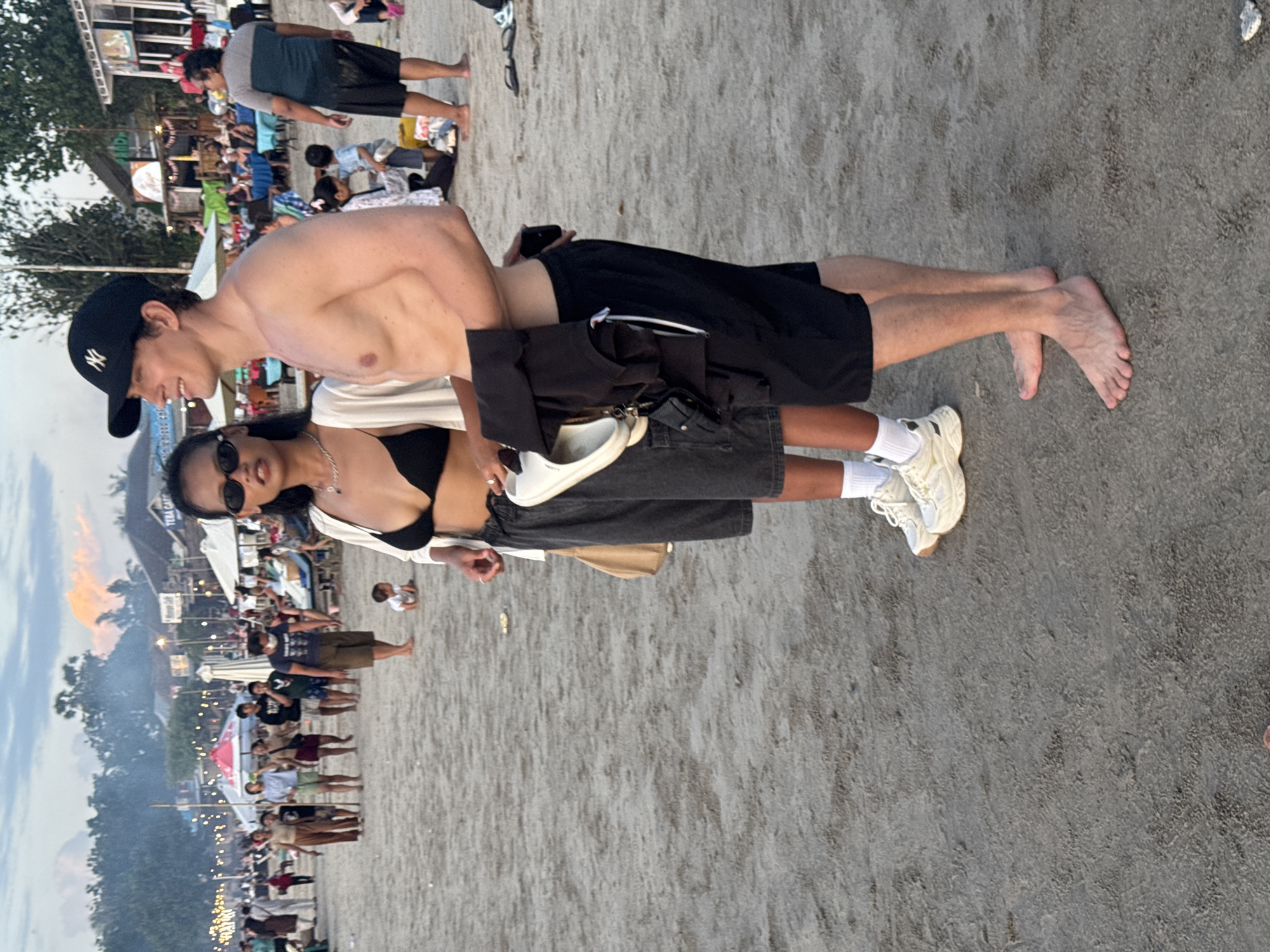 On a beach in Kuta Selatan, Indonesia, two individuals, a man and a woman, are the focal point of the scene. The man is shirtless, wearing a black cap and black shorts. The woman is wearing sunglasses, a black bikini top, a white shirt, and gray shorts. They are standing close together. The background shows a crowded beach with other people, trees, and lit-up structures. The sky is visible in the background, suggesting it's either sunrise or sunset.