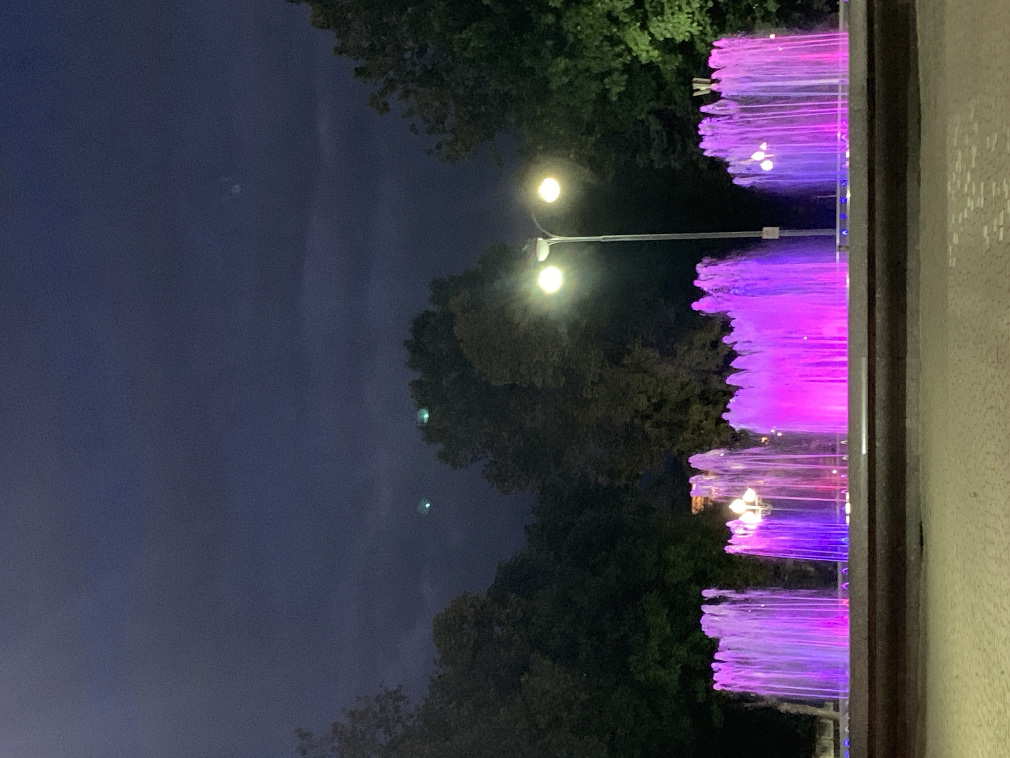 At night in an outdoor urban setting, a row of illuminated fountains is the central focus. The water jets are lit with vibrant purple and pink lights, creating a colorful spectacle against the dark sky. Trees line the background, their silhouettes visible in the dim light. A street lamp with two globes stands to the right of the fountains, its warm yellow light illuminating the immediate surroundings. The ground in the foreground appears to be paved, with a textured surface. The sky is a deep, cloudy blue, with a few faint, indistinct green lights visible high up, possibly aircraft or atmospheric phenomena. The overall scene suggests a public park or square that is active in the evening.