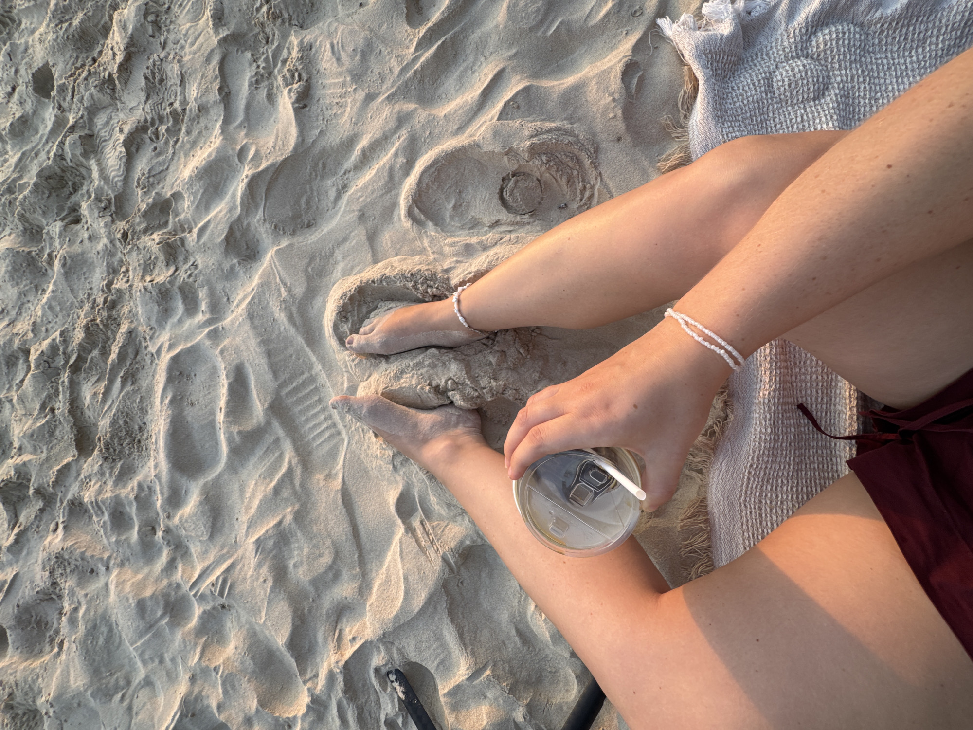 In Karon, Thailand, a person sits on a sandy beach, illuminated by the warm glow of sunset. The sand is textured with ripples and footprints, suggesting a well-trafficked area. The individual's legs, adorned with white beaded anklets and bracelets, are crossed. They hold a transparent cup with a white lid and straw, appearing to contain a light-colored beverage, possibly coffee or tea. The person is wearing dark red shorts. A light-colored woven towel lies beneath their legs. The scene captures a moment of relaxation on a tranquil beach.