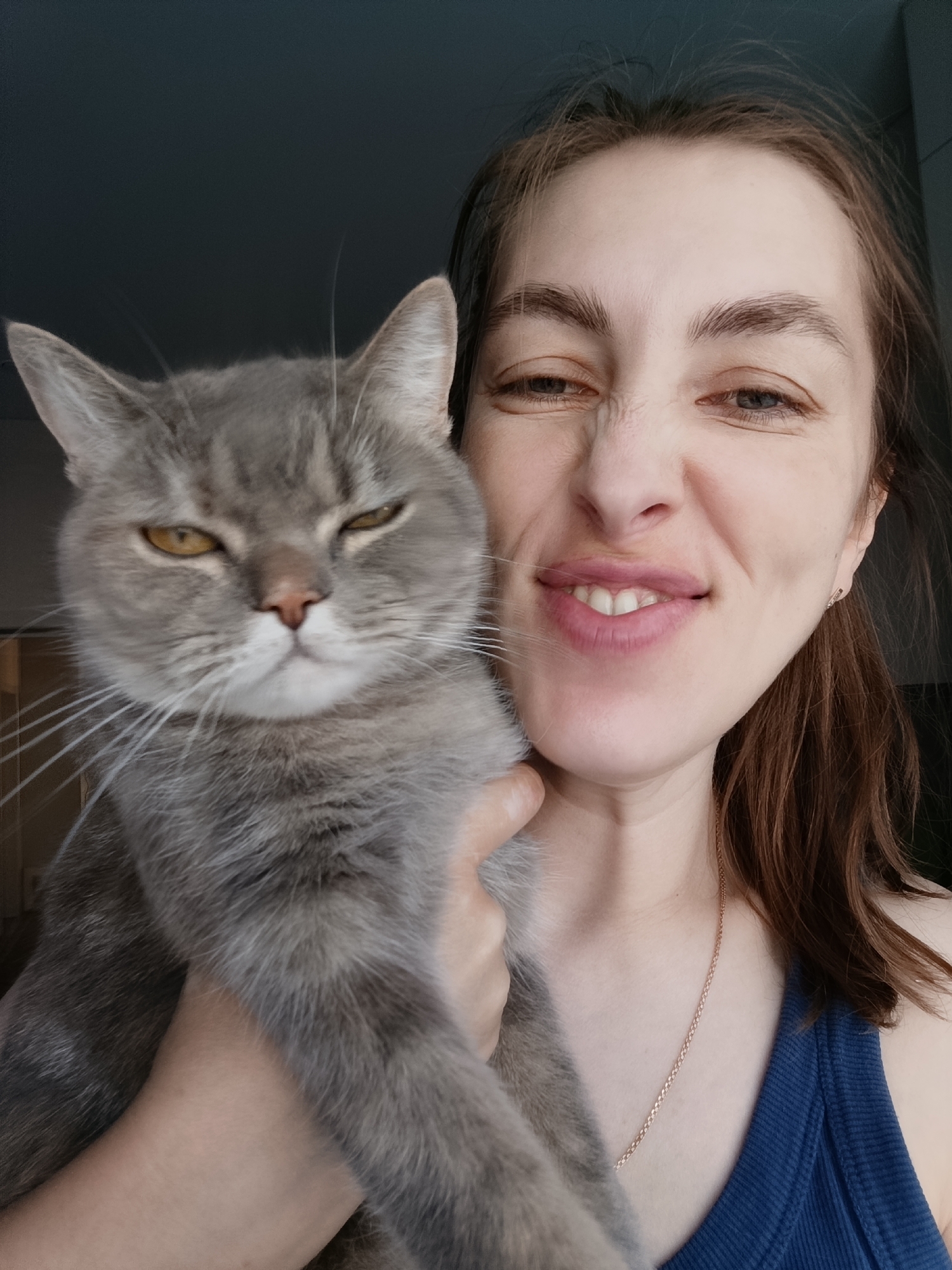 A woman and a gray cat are the primary subjects of this indoor selfie. The woman is holding the cat, positioning it close to her face. The woman is smiling with a slight grimace, possibly due to holding the cat, and the cat appears to be squinting. The woman is wearing a blue tank top and a gold necklace. The location is Chaikovsky, Russia.