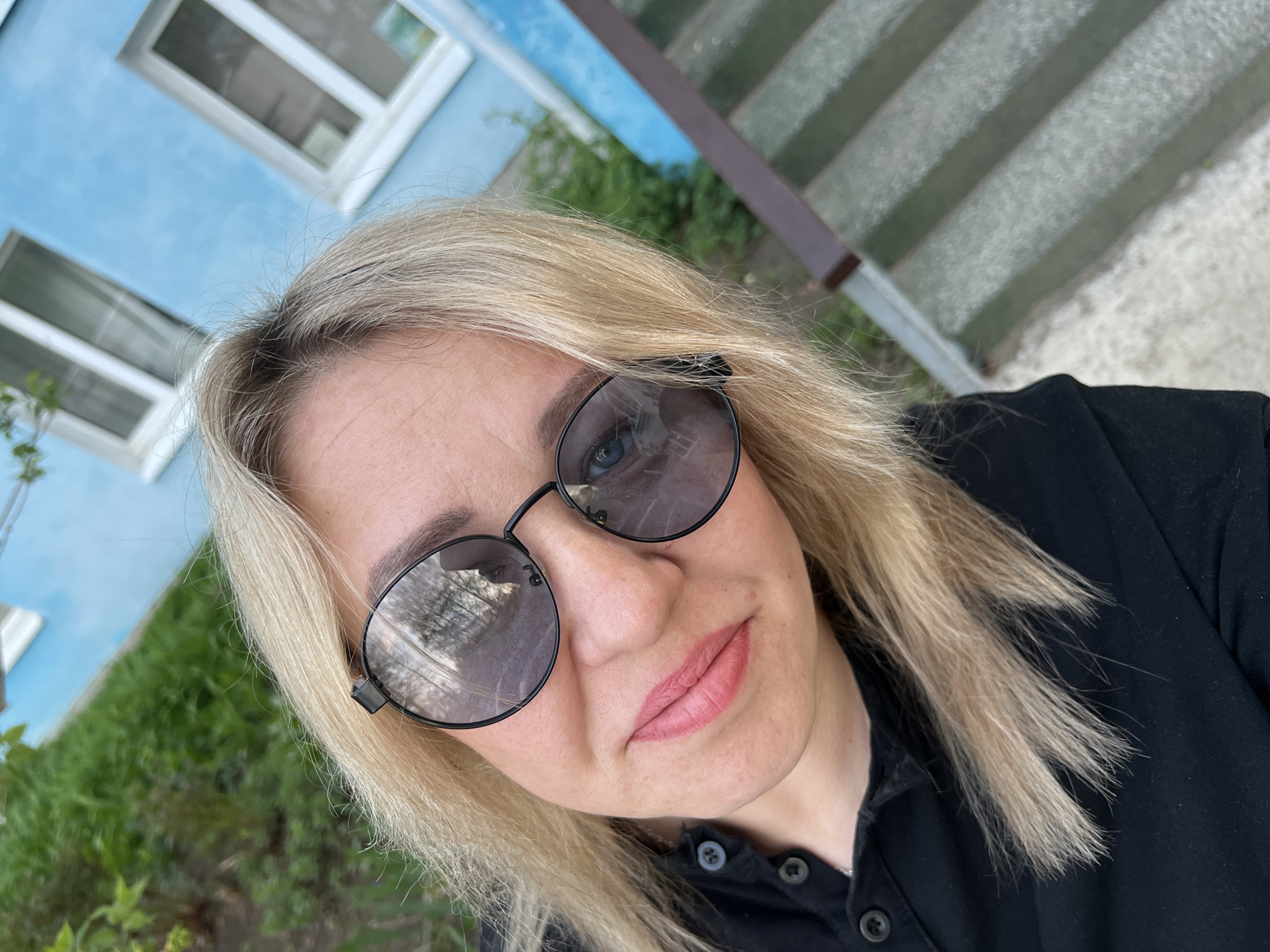 A fair-skinned, blonde woman wearing sunglasses and a black button-up shirt is taking a selfie. Behind her is a light-blue building with white-framed windows. Adjacent to the building is a grey concrete structure with a brown railing. Green foliage is partially visible at the bottom of the frame. Based on the location information, the scene is in Chaikovsky, Russia.