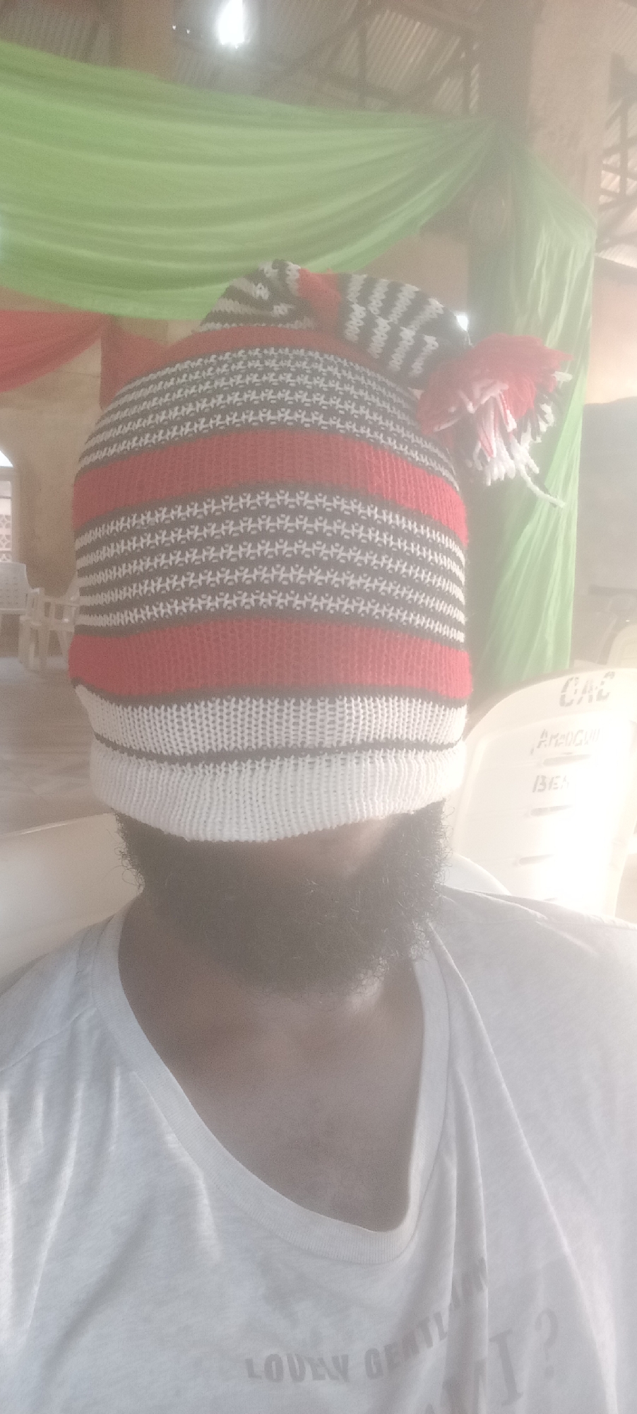 A man with a full beard wears a striped knit cap pulled down over his eyes. The cap is primarily red, white, and gray, with a pom-pom on top. He is wearing a white t-shirt, and the setting appears to be indoors with green and red fabric draping in the background. The location is potentially Bende, Nigeria, suggested by context. White plastic chairs are visible in the background.