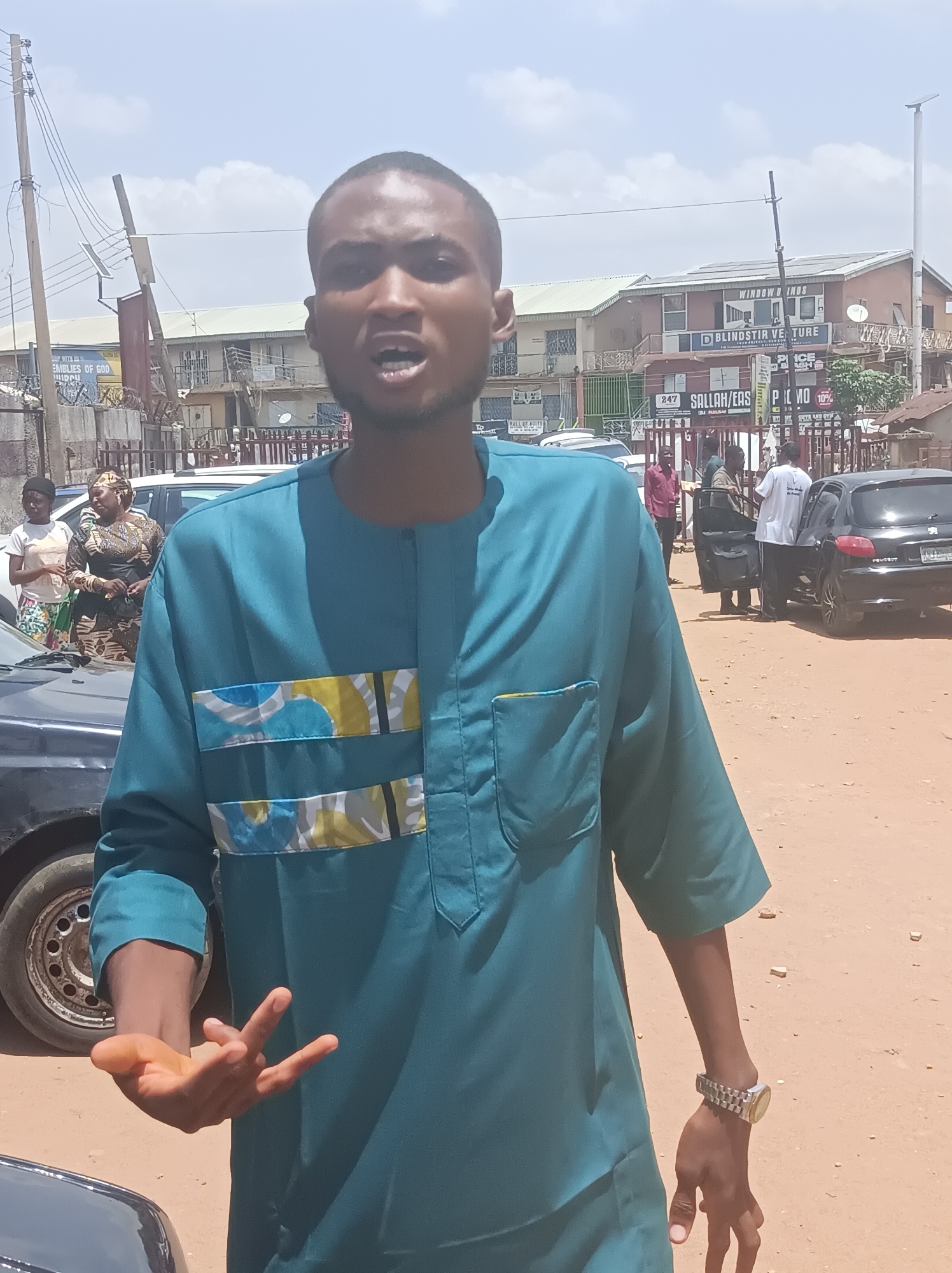 A man in a teal shirt with patterned blue, yellow, and white accents stands outdoors on a dirt ground. He is gesturing with his left hand and wearing a silver wristwatch on his right wrist. In the background are several people, parked cars, and buildings. A sign reads "BLINDSTIR VENTURE" and "SALLAH/EAS." The location is Dadi Riba, Nigeria.
