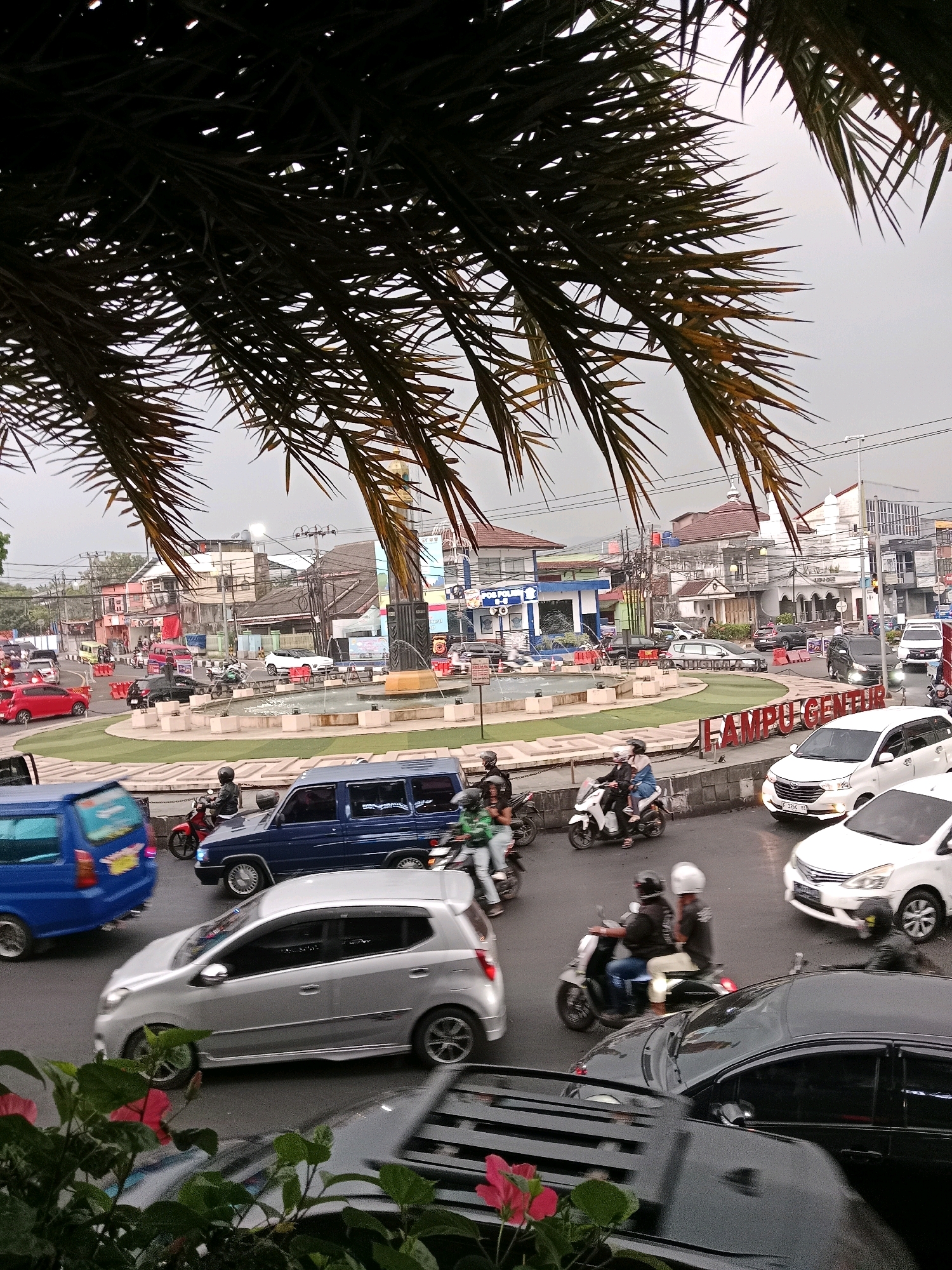 The image captures a busy intersection in Cianjur, Indonesia, with a circular fountain at its center. A variety of vehicles, including cars, motorcycles, and a blue van, navigate the roundabout under an overcast sky. Several palm fronds frame the top of the scene, suggesting the presence of tropical vegetation. Buildings, some with signs in Indonesian, line the surrounding streets, indicating a commercial or residential area. People are visible on motorcycles, some riding with passengers, while others are driving cars or walking. The overall impression is one of urban activity on a typical day.