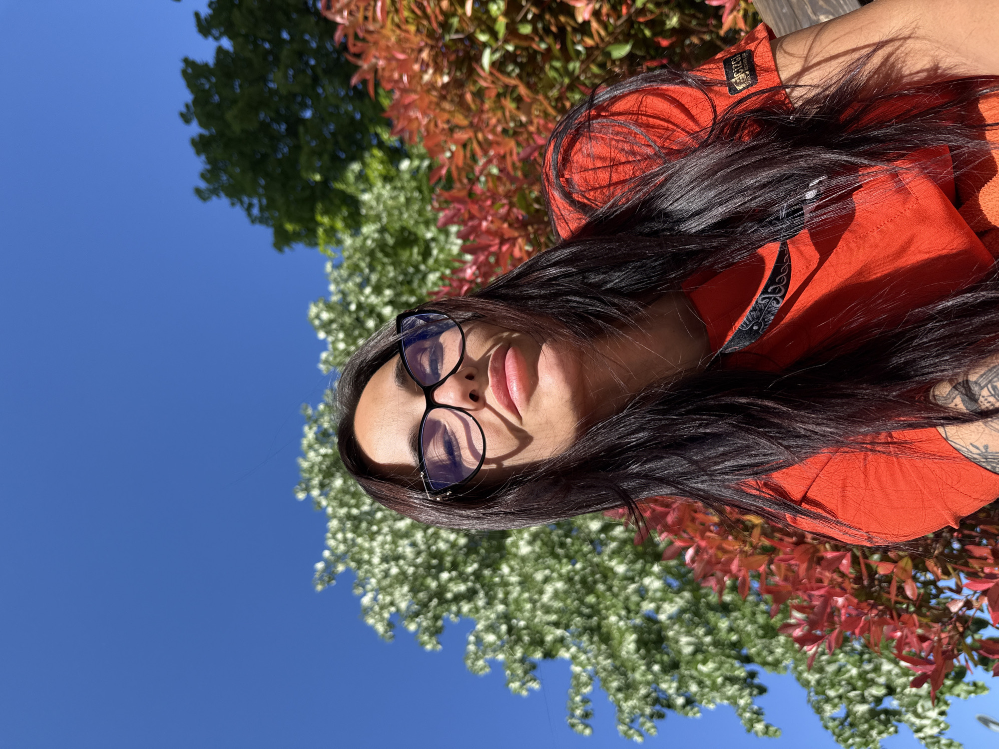 A woman with long dark hair and glasses is lying down, possibly outdoors, with a bright blue sky, green foliage, and reddish-orange foliage in the background. The location may be Pleven, Bulgaria. The woman is wearing an orange garment. A tattoo is visible on her right arm.