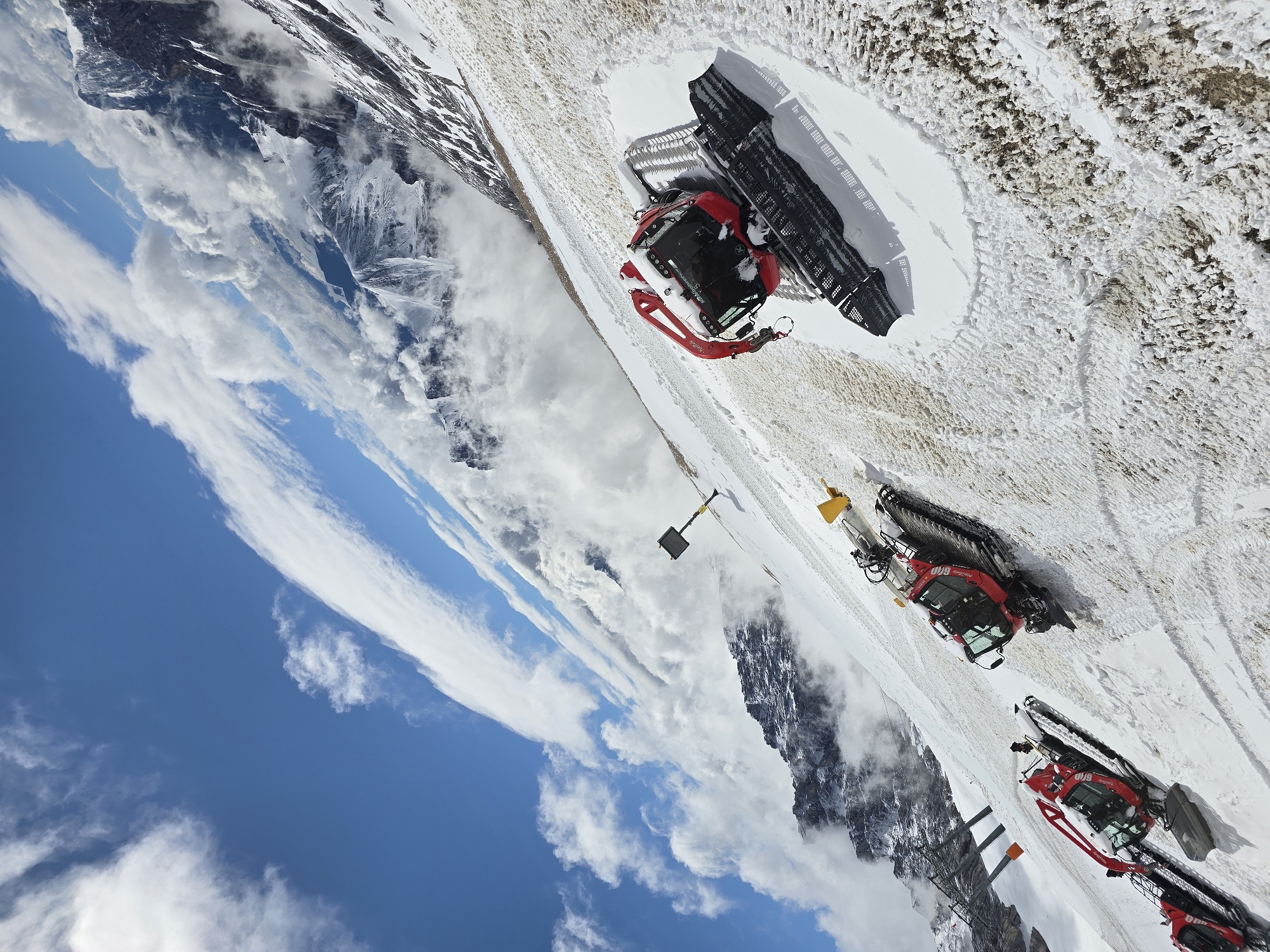 This is a high-angle, wide shot of four red snow groomer vehicles on a snowy mountainside, possibly in Garmisch-Partenkirchen, Germany, under a bright blue sky with scattered white clouds. The scene is tilted, giving a sense of steepness. The snow is a mix of fresh white powder and darker, compacted snow, with visible tracks from the vehicles. In the background, rugged, snow-covered mountains rise up, partially obscured by clouds. A yellow post with a square black sign is visible on the slope. The snow groomers are parked on the slope, with the closest one in the foreground exhibiting detailed tracks in the snow around its treads.