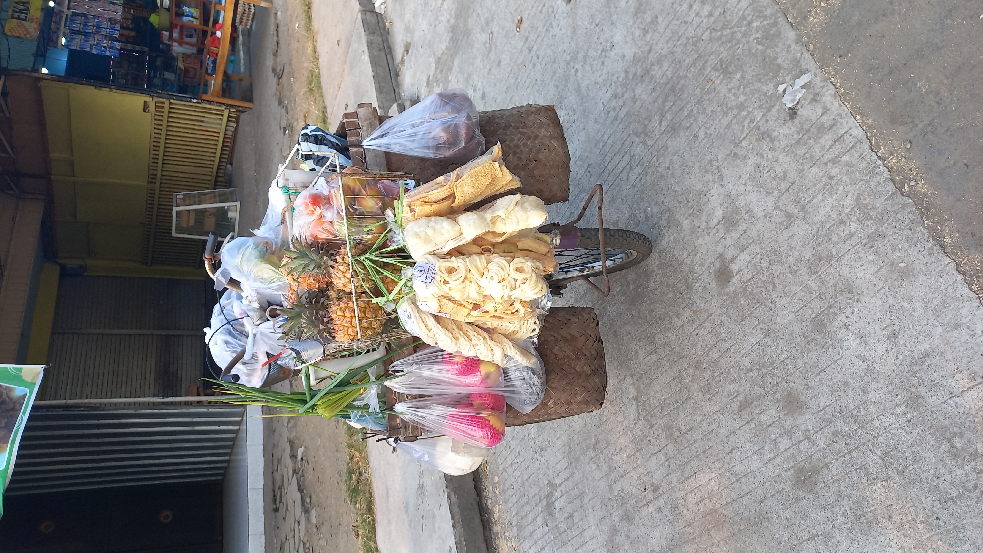The image depicts a street scene in Mangunsari, Indonesia, focusing on a bicycle laden with goods. The bicycle is positioned on a concrete path, with a store facade visible in the background, characterized by shuttered storefronts and a yellow awning. The bicycle's cargo includes an assortment of fruits, primarily pineapples and some red fruits, as well as packaged snacks and what appear to be vegetables. The fruits are loosely packed in baskets and plastic bags, while the snacks are in clear plastic packaging. The bicycle itself is a simple model with a metal frame and a wire basket at the front. The ground is concrete, showing signs of wear and some debris. The overall impression is a vendor's mobile cart, ready for business.