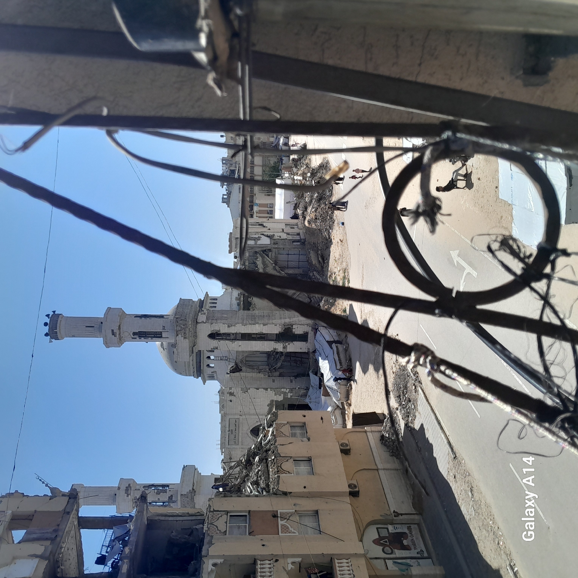 The image displays a destructed urban landscape in Khan Yunis, Palestinian Territory, viewed from an elevated position through a dark, blurred metal grille. In the mid-ground, the severely damaged Al-Farouk Mosque stands prominently, featuring a partially intact minaret and a heavily compromised dome. Extensive structural breaches are visible across its walls, with large piles of debris surrounding its base.
To the immediate left, a multi-story building exhibits catastrophic damage, its upper floors collapsed into rubble and exposing internal structures. The lower section of this building shows windows, a balcony, and faded graffiti on the wall, including a red triangle symbol and "Squid Game" text, along with "YASEEN GAMES".
An asphalt road traverses the mid-ground, bearing a white arrow painted on its surface, indicating a right turn. This road is flanked by sandy ground and scattered construction debris. Several individuals are present: a lone figure sits on a black chair near the road's right edge, and multiple small groups of people, including adults and children, walk along the road and adjacent sandy areas. In the background, beyond the mosque and road, additional damaged buildings are visible, interspersed with white tarpaulin tents, suggesting temporary shelters. The scene is illuminated by clear daylight under a bright blue sky. Overhead, thin black lines, consistent with electrical or communication cables, cross the upper frame. A "Galaxy A14" watermark is present at the bottom left.