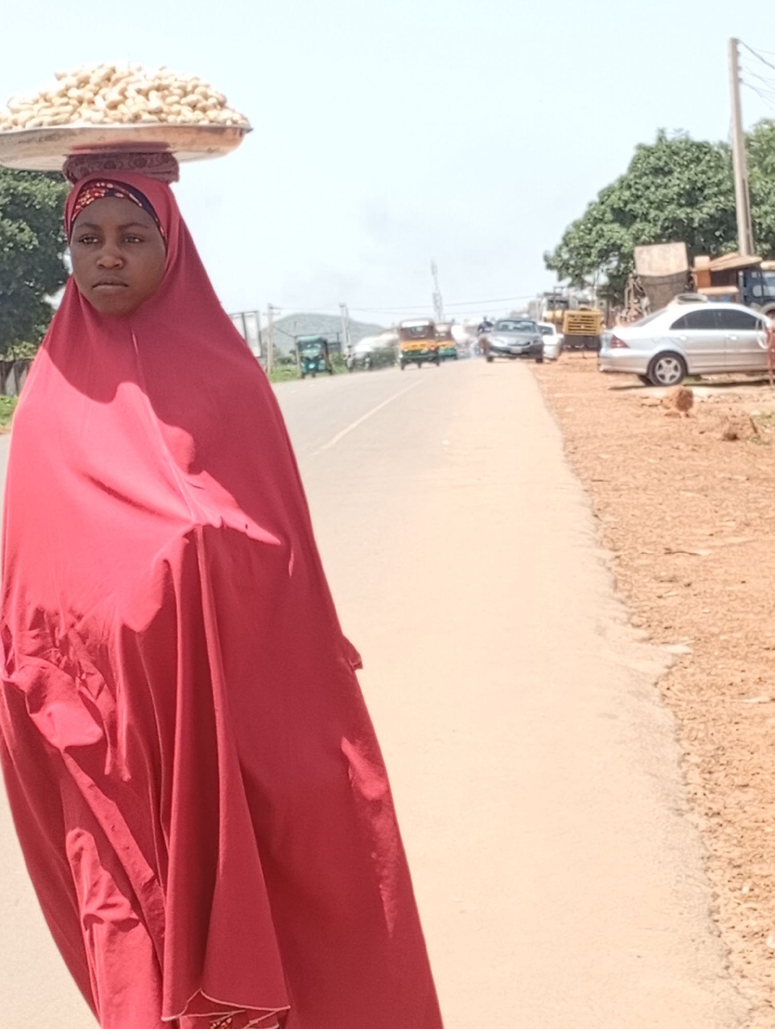A young woman, likely a vendor, walks on the side of a paved road in Rafin Par, Nigeria. She is wearing a vibrant red headscarf and a matching flowing garment that covers her from head to toe. Balanced precariously on her head is a large metal tray filled with what appear to be groundnuts. Her expression is neutral as she walks, her gaze directed forward. The road stretches into the distance, with a few vehicles visible, including what looks like a silver sedan, a truck, and some three-wheeled vehicles. The surrounding environment is dry and dusty, with sparse vegetation and scattered structures. The sky is overcast, suggesting a hazy day. The scene captures a moment of daily commerce and life on a Nigerian roadside.