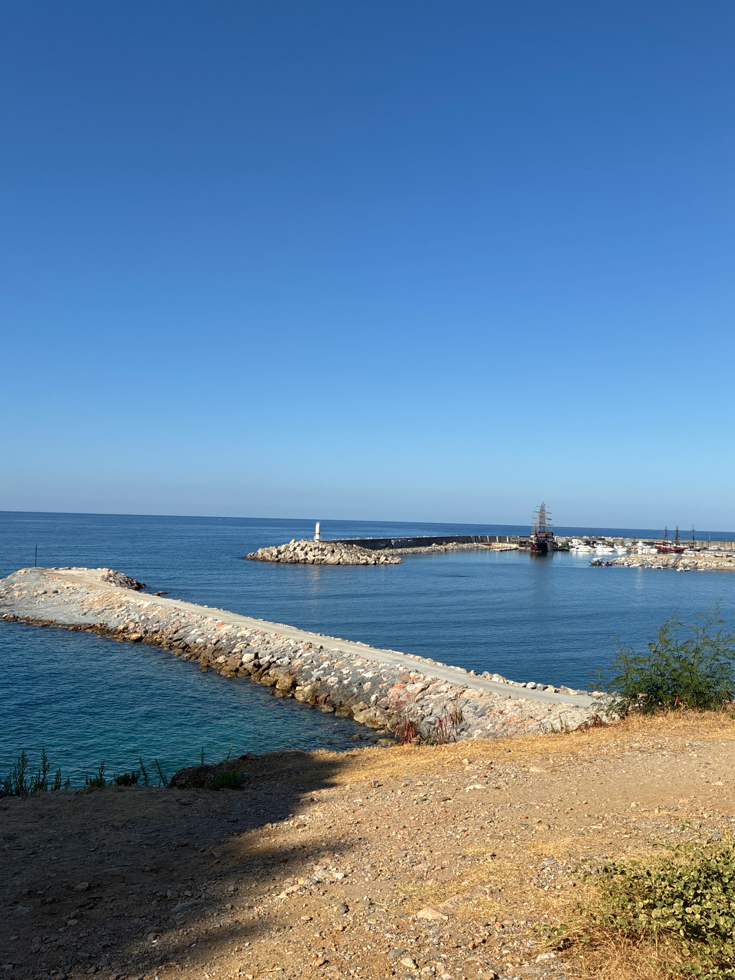 This is a photograph of the harbor entrance in Alanya, Turkey, on a clear, sunny day with a bright blue sky. A long, rocky breakwater extends into the blue sea from the left of the frame. A second, wider breakwater, also constructed of rocks, forms an arc and encloses a harbor. On this second breakwater, a small white lighthouse with a red top is visible. Within the harbor, several boats are docked, including a large, dark-colored ship with multiple masts, resembling a pirate ship. Another similar vessel can be seen further in the harbor. The foreground of the image consists of dry, dusty ground with scattered rocks and sparse, dry vegetation, suggesting a warm climate. Some green bushes are visible on the right side of the frame. The overall impression is a coastal scene of a Mediterranean harbor.