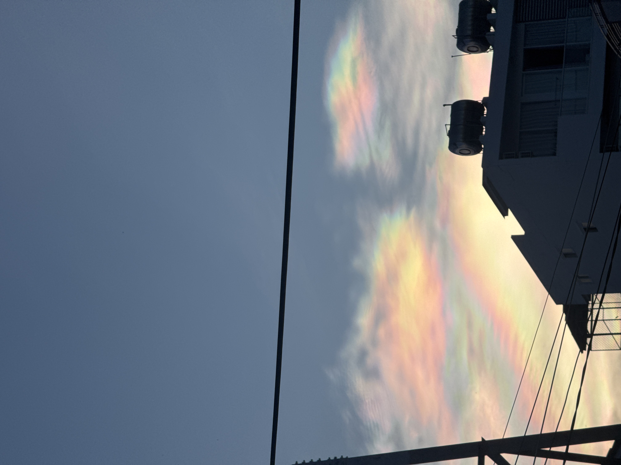 The image, taken in or near Bac Nha Trang Ward, Vietnam, captures a segment of the sky, power lines, and a building. The left portion of the image shows a clear, pale blue sky. A prominent vertical power line bisects the image. To the right of the power line, iridescent clouds display a spectrum of colors – pink, yellow, green, and blue – suggesting the occurrence of cloud iridescence. A building structure occupies the right side of the image. The silhouette of the building is visible, featuring two water tanks on the roof. The photograph is taken from a low angle, looking upwards, with power lines running across the bottom portion.