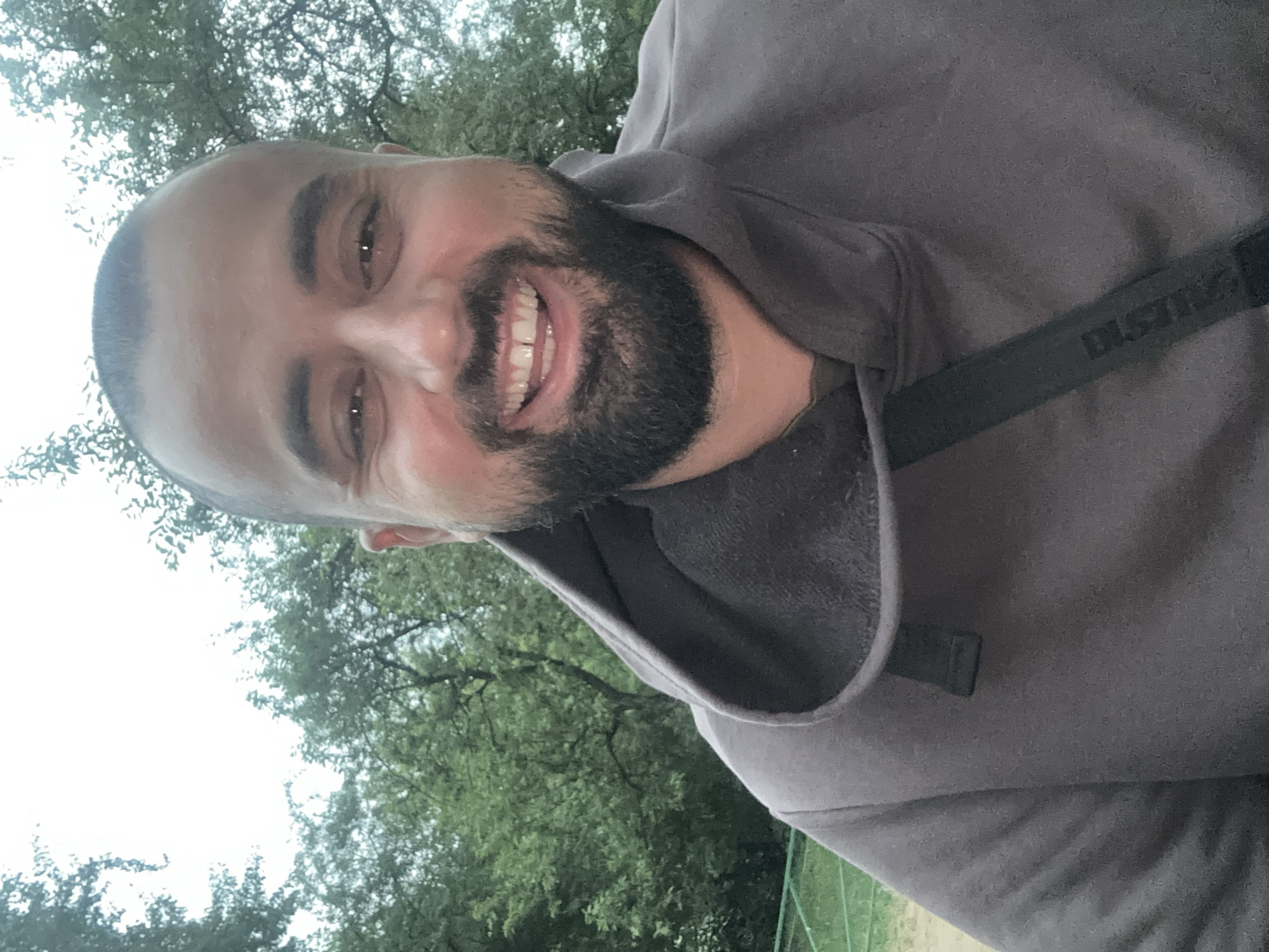 The image is a close-up, selfie-style shot of a man with a shaved head and a beard. He is smiling broadly, showing his teeth. He is wearing a dark grey hooded sweatshirt with the hood up. A black strap with some visible lettering is visible across his chest. The background consists of out-of-focus green trees and a bright, overcast sky. Based on the provided information, the location is the city of Lytkarino, Russia.