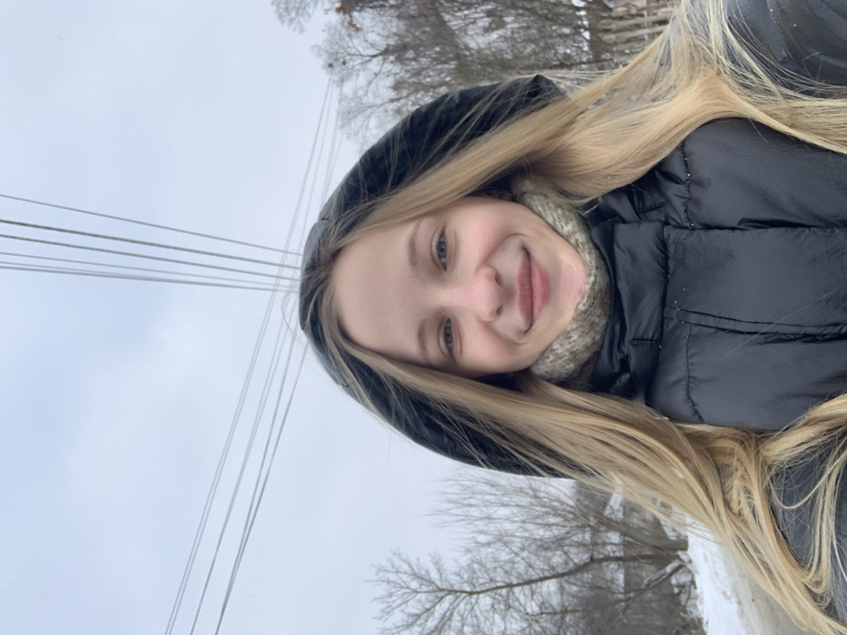 A young female with long blonde hair, blue eyes, and a smiling expression is captured in a self-portrait taken outdoors in Bohuslav, Ukraine. She is wearing a black hooded puffer jacket, with the hood up, and a textured greenish-grey neckwear item. The background consists of an overcast, light grey sky dominating the upper portion of the frame. Multiple parallel utility lines stretch horizontally and diagonally across the sky. Bare deciduous trees are visible on both sides of the subject, extending into the middle ground. Snow partially covers the ground on the left, and distant structures or fences are faintly discernible through the trees, consistent with a winter setting.