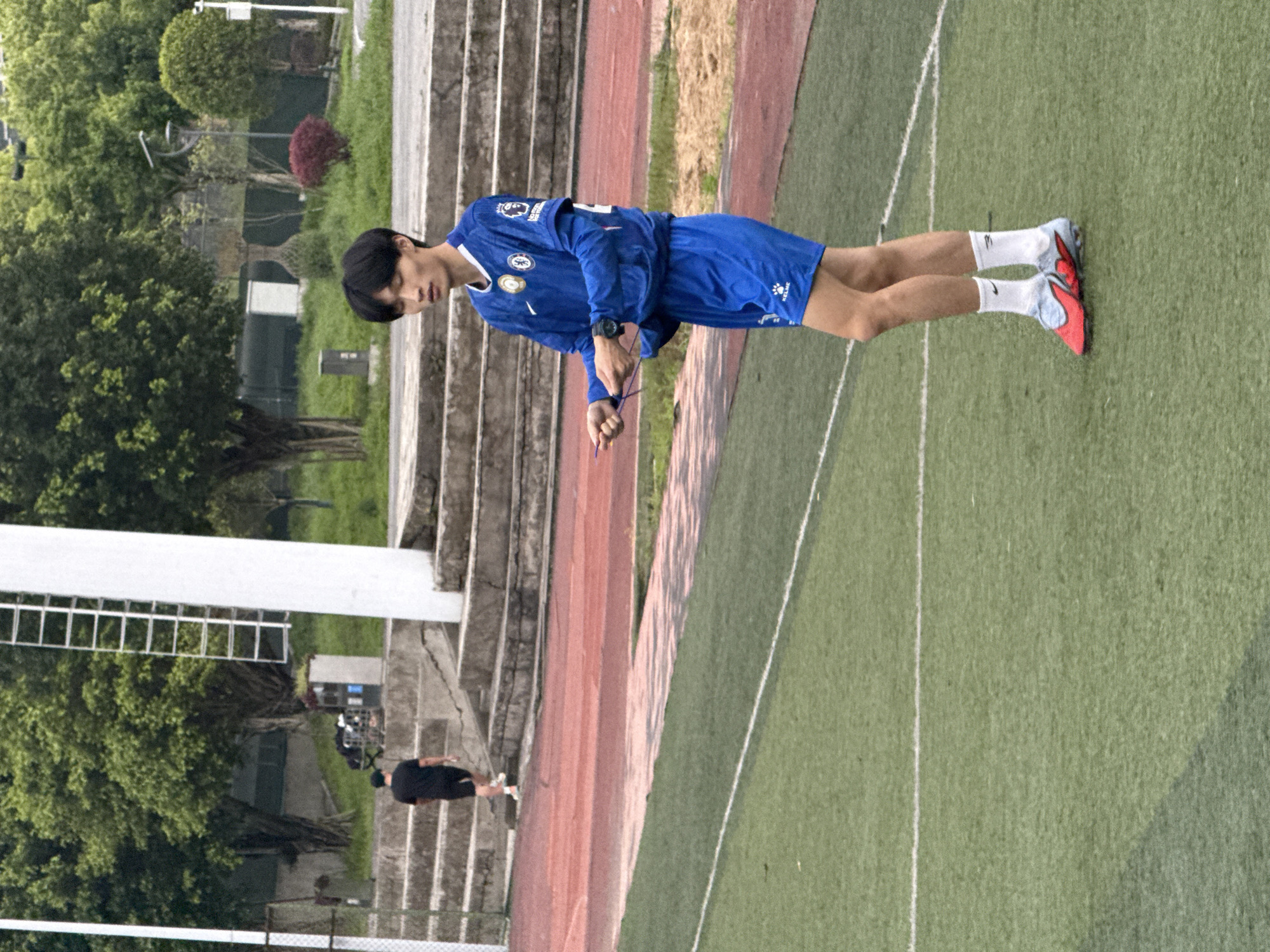 A young adult male wearing a blue Chelsea soccer uniform stands on a green grass field with white lines. He is tying a purple shoelace. He has black hair and is wearing white socks and red and blue soccer cleats. To the left of the field is a red track, and next to that, concrete steps leading up to a grassy area with trees and a white pole structure. Another individual dressed in black is walking up the steps in the background. The location is the city of Jiangjin, China.